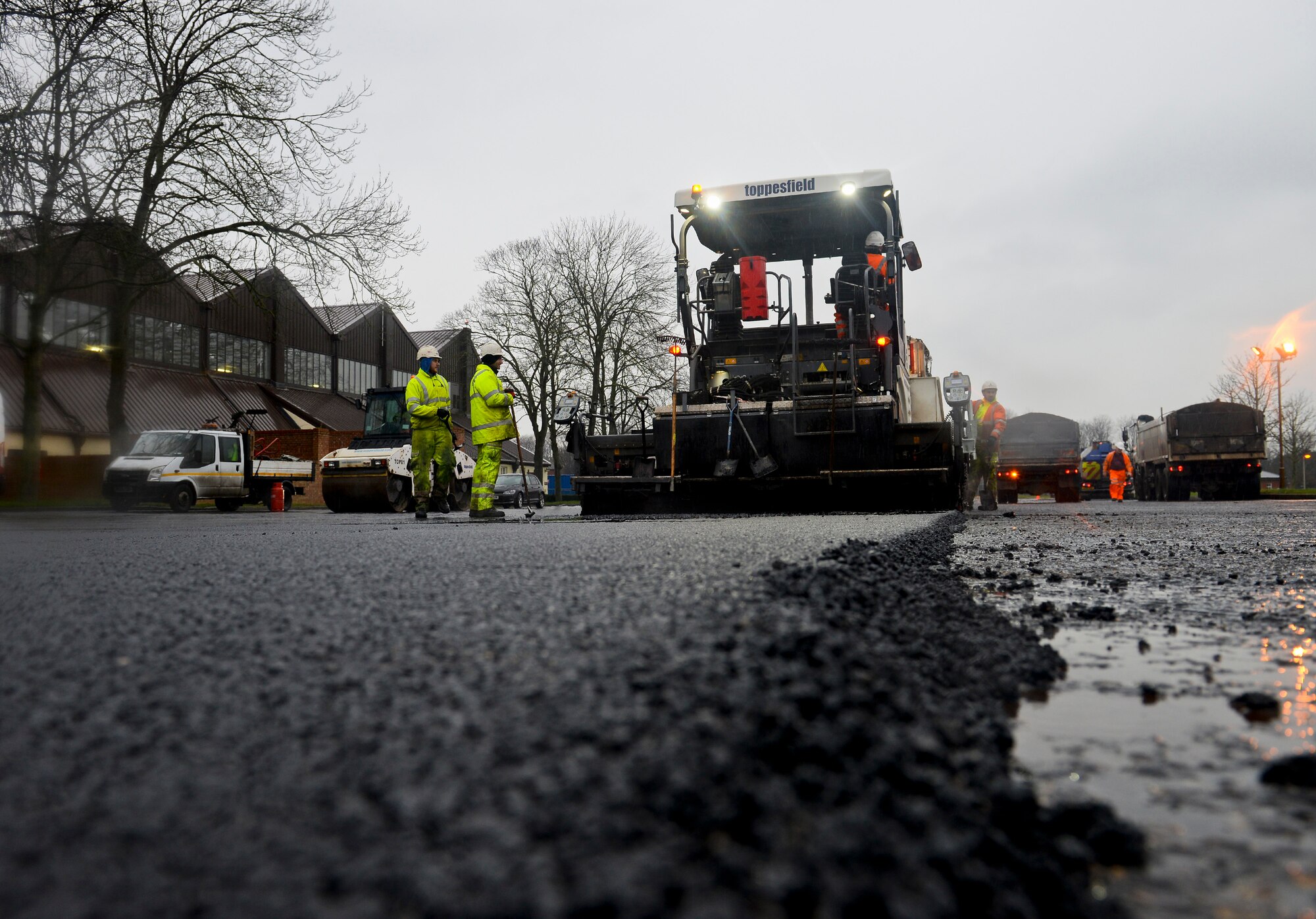 A contracted construction worker operates an asphalt paver to lay hot asphalt as other contracted construction workers spread hot tarmac over the 100th Logistics Readiness Squadron Vehicle Operations yard Jan. 22, 2016, on RAF Mildenhall, England. The Vehicle Operations yard was resurfaced to maintain a serviceable infrastructure. (U.S. Air Force photo by Staff Sgt. Micaiah Anthony/Released)