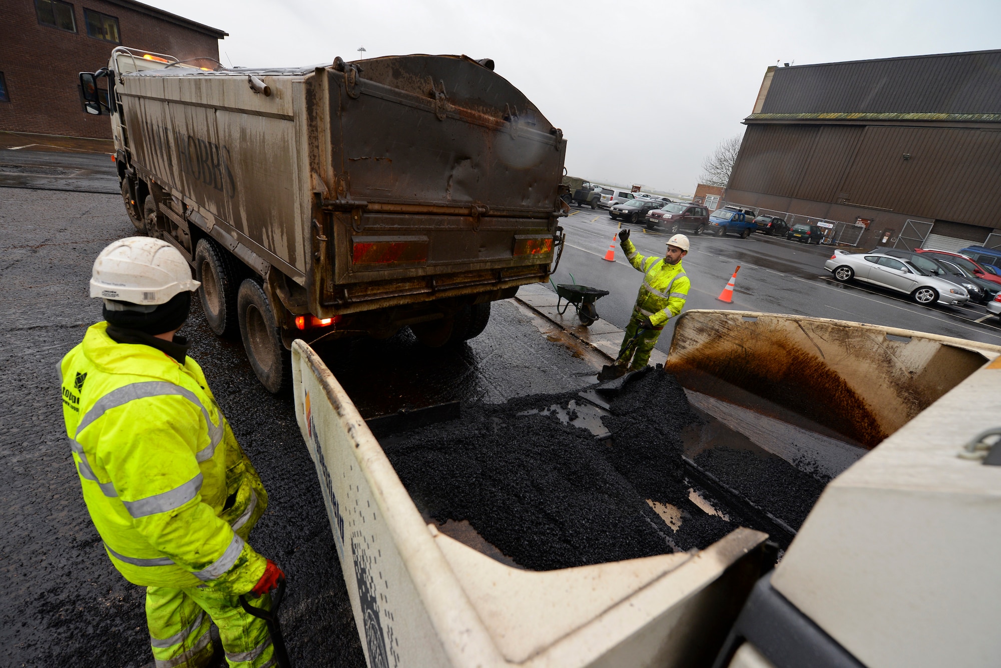 Contracted construction workers guide a dump truck to an asphalt paver in the 100th Logistics Readiness Squadron Vehicle Operations yard Jan. 22, 2016, on RAF Mildenhall, England. The dump trucks have tarps fitted over them to prevent heat from the hot tarmac from escaping.  Asphalt is heated up to 392 degrees Fahrenheit before it is loaded into the truck. (U.S. Air Force photo by Staff Sgt. Micaiah Anthony/Released)