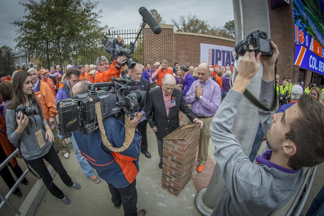 U.S. Army Col. (retired) and Bataan Death March survivor Ben Skardon is surrounded by media and well-wishers - including Clemson University President James Clements (right) - as he reads the plaque at the base of the Memorial Stadium flag pole that was dedicated to him at a ceremony Nov. 21, 2015. (U.S. Army photo by Staff Sgt. Ken Scar)