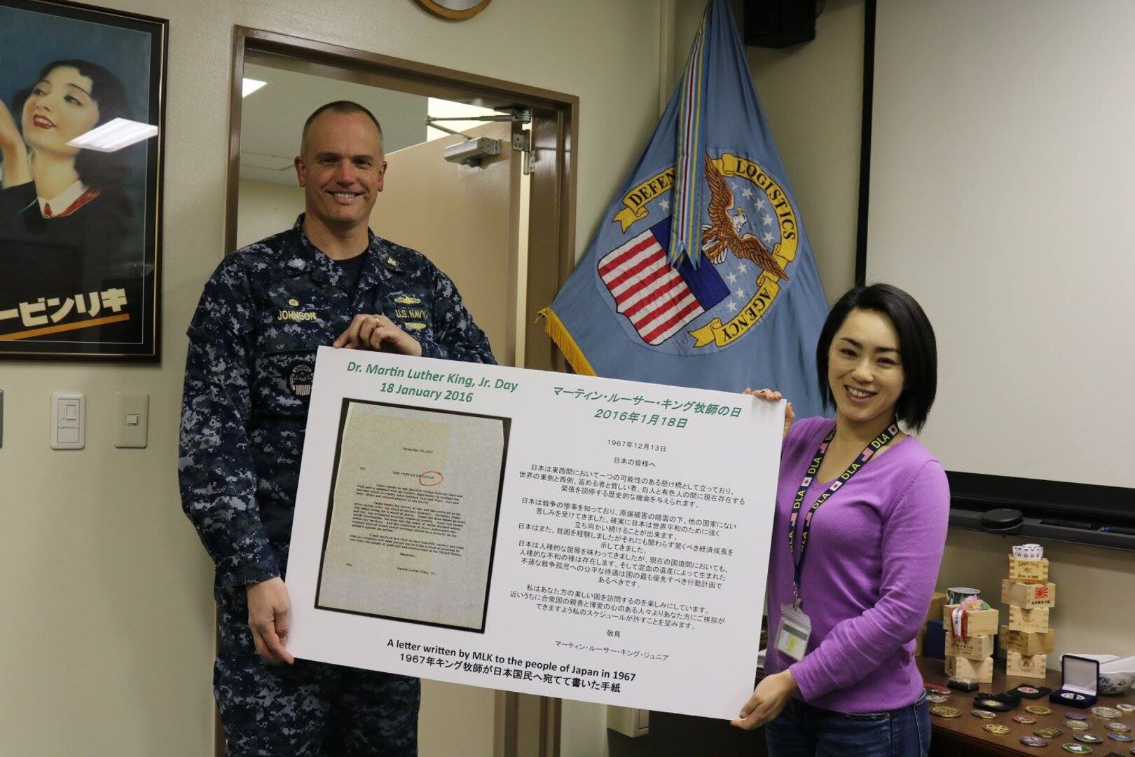 Navy Cmdr. Brian Johnson, DLA Distribution Yokosuka, Japan, commander, and Tomo Oobuko, the distribution center’s cultural champion, display a board highlighting Dr. Martin Luther King Jr. Day. The poster board shows a copy of the letter Dr. King wrote to the Japanese people, and the Kanji translation.  
 
