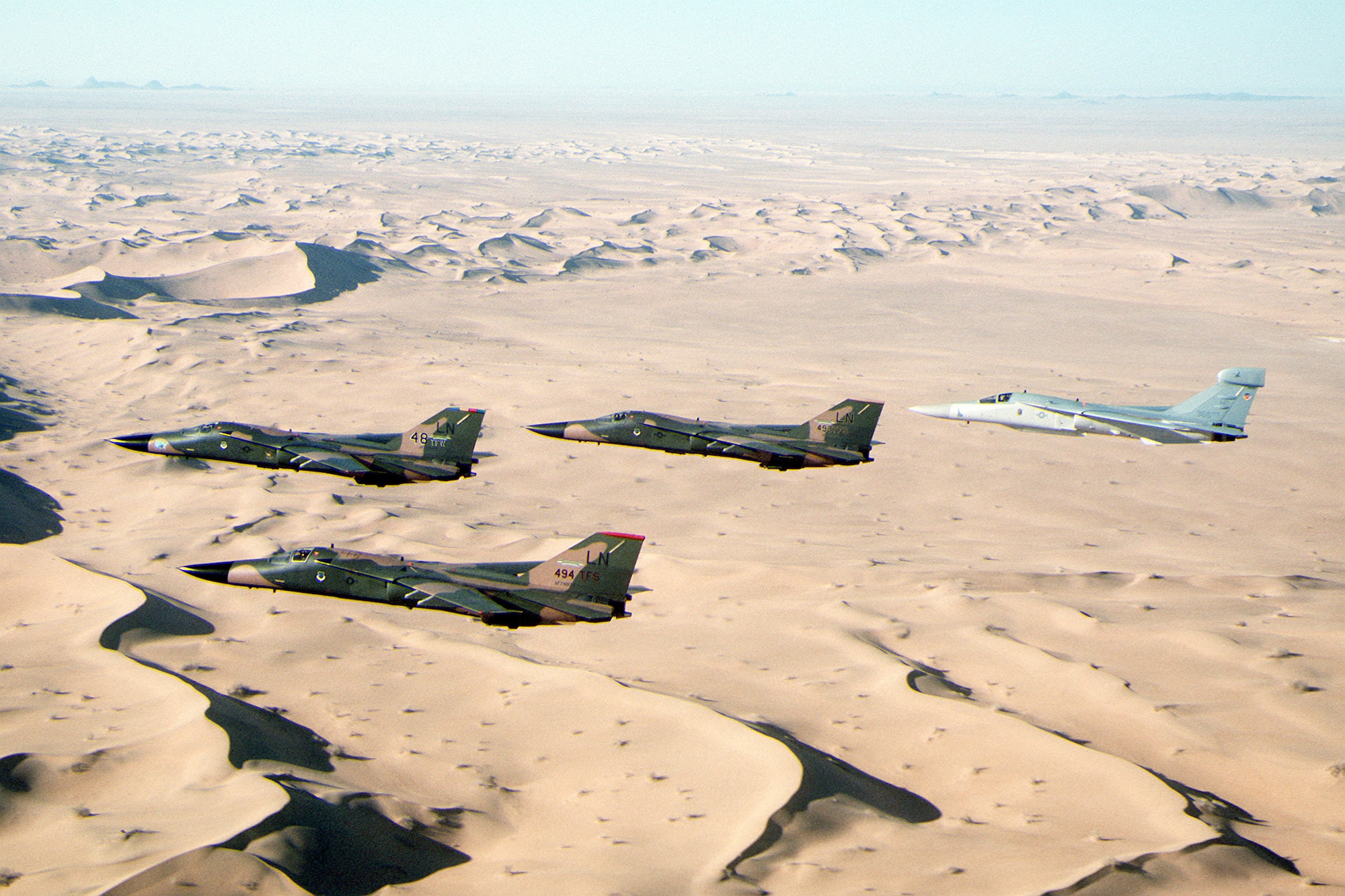 F-111F aircraft and an EF-111A Raven, rear, fly in formation over the desert during Operation Desert Shield. The aircraft are assigned to the 48th Tactical Fighter Wing. (Courtesy photo)