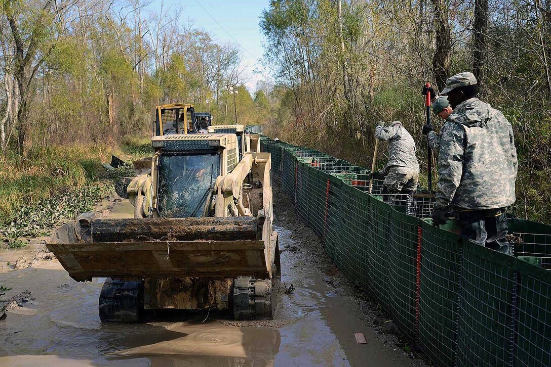 A soldier operates a skid loader through muddy water while fellow soldiers fill barriers that are being used to construct a levee on Avoca Island, La., Jan. 11, 2016. Louisiana National Guard photo by Army Spc. Joshua Barnett
