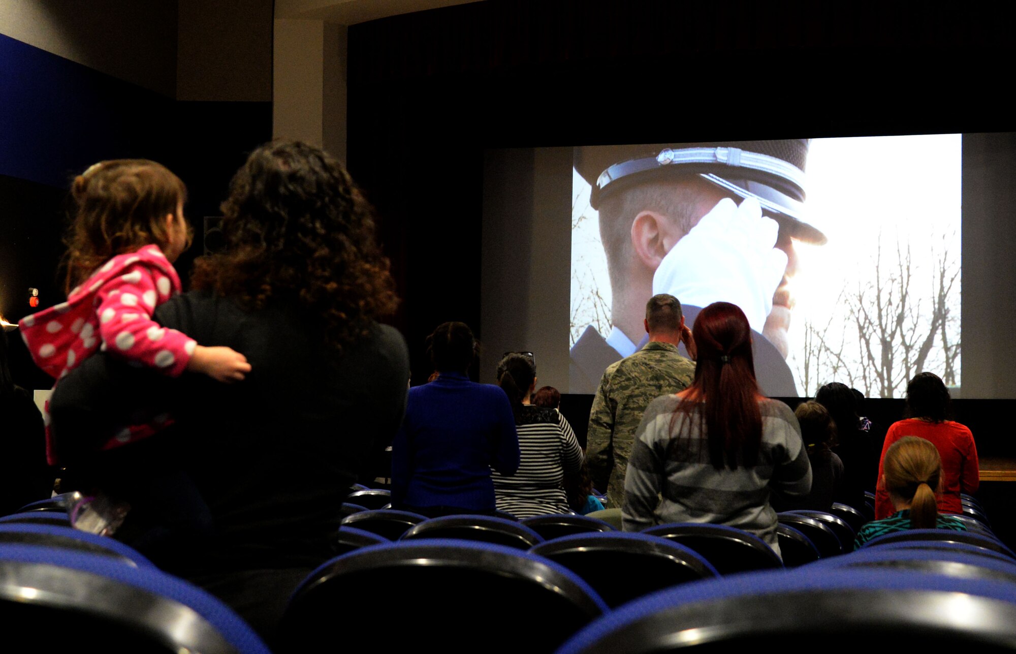 Attendees of the Hearts Apart event stand for the playing of the national anthem before a viewing of The Peanuts Movie Jan. 14, 2016, at the base theater on RAF Mildenhall, England. Team Mildenhall Spouses Club provided snacks, goodie bags and a craft table to encourage a kid-friendly theater environment. The evening’s events were to help bring families closer together and develop a sense of community while their loved ones are deployed.  (U.S. Air Force photo by Airman 1st Class Justine Rho/Released)