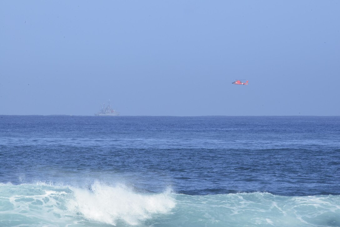 A crew aboard an MH-65 Dolphin helicopter flies a search pattern over the North Shore of Oahu, Jan. 18, 2016, during efforts to locate 12 missing Marines. The helicopter is assigned to Coast Guard Air Station Barbers Point, Hawaii. The USS Paul Hamilton, back left, also assisted in the search. U.S. Coast Guard photo by Petty Officer 1st Class Levi Read