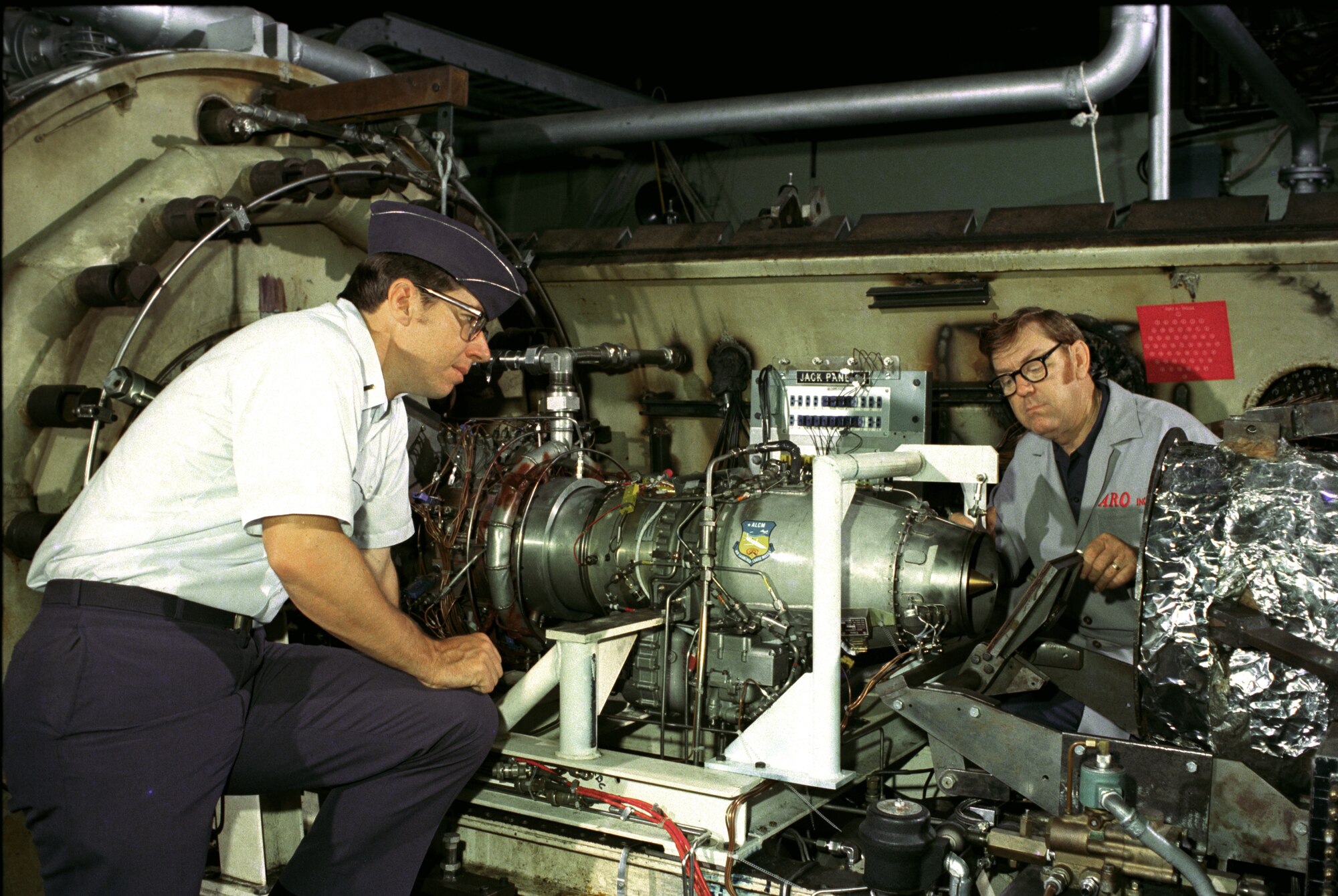 AEDC personnel check a quick-opening at the F107 turbofan engine's exhaust nozzle. The door was opened as air was supplied to the engine's inlet to simulate an in-flight start. The F107 engine powers Air-Launch Cruise Missiles, such as the AGM-86C used by the B-52 bomber. (AEDC Photo-1975)