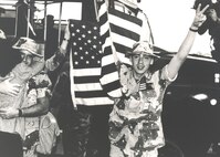 SrA Kevin D. Schwinn waves the American flag and flashes a victorysign following his return from Desert Storm with other members of the15th Security Police Squadron. At left, getting a big hug, is AlCPatrick S. Waters. (Courtesy Photo)