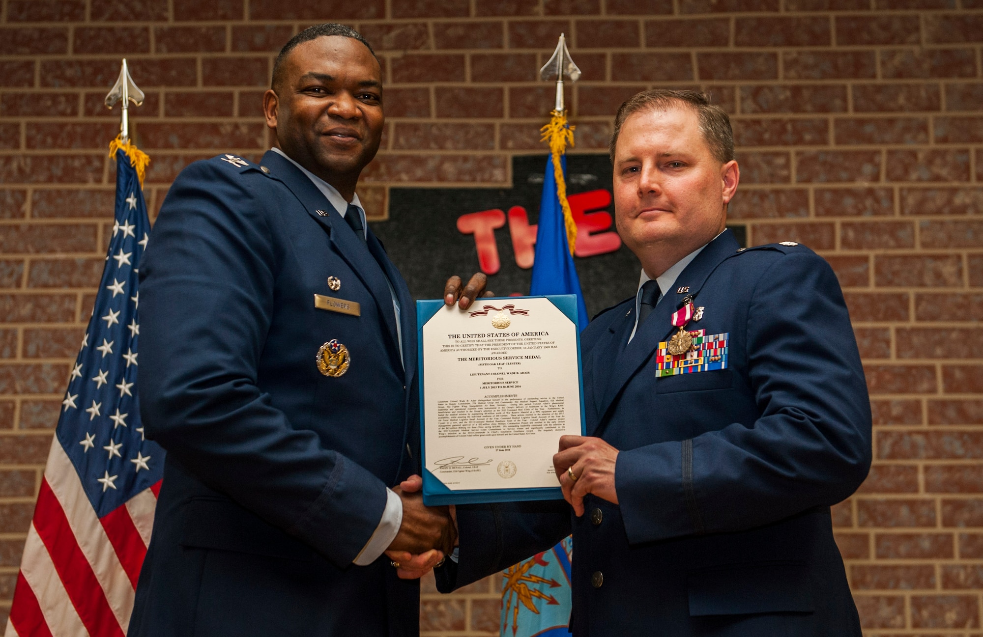 U.S. Air Force Col. Alfred Flowers, 52nd Medical Group commander, left, and U.S. Air Force Lt. Col. Wade Adair, outgoing 52nd Medical Support Squadron commander, hold up a certificate of the Meritorious Service Medal during the 52nd MDSS change of command ceremony in the Brickhouse on Spangdahlem Air Base, Germany, June 30, 2016. Adair received a Meritorious Service Medal in recognition of his service of leading the 52nd MDSS. (U.S. Air Force photo by Airman 1st Class Timothy Kim/Released)