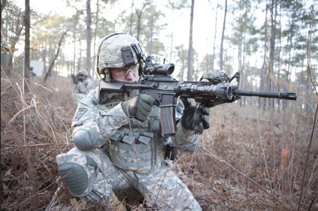 First lieutenant Officer wearing ACUs aiming Carbine outside during the day in the woods, fellow soldier in background.