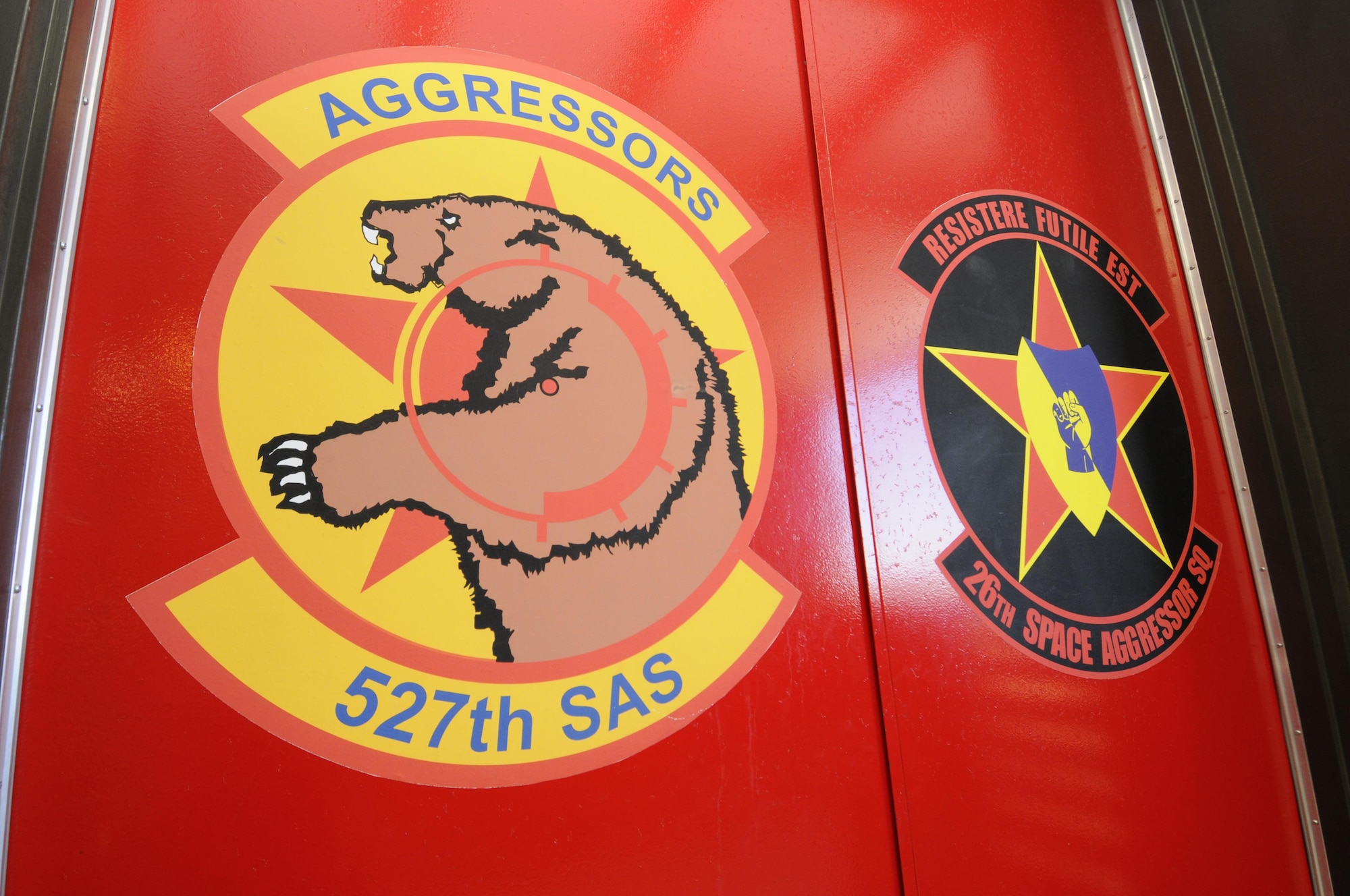 The 527th Space Aggressors Squadron and 26th Space Aggressors Squadron emblems are painted side by side on doors inside the Space Aggressors’ facility at Schriever Air Force Base, Colorado. Currently, the Space Aggressors replicate live GPS and SATCOM electronic attack for training audiences across the globe.  (U.S. Air Force photo/2nd Lt. Darren Domingo)
