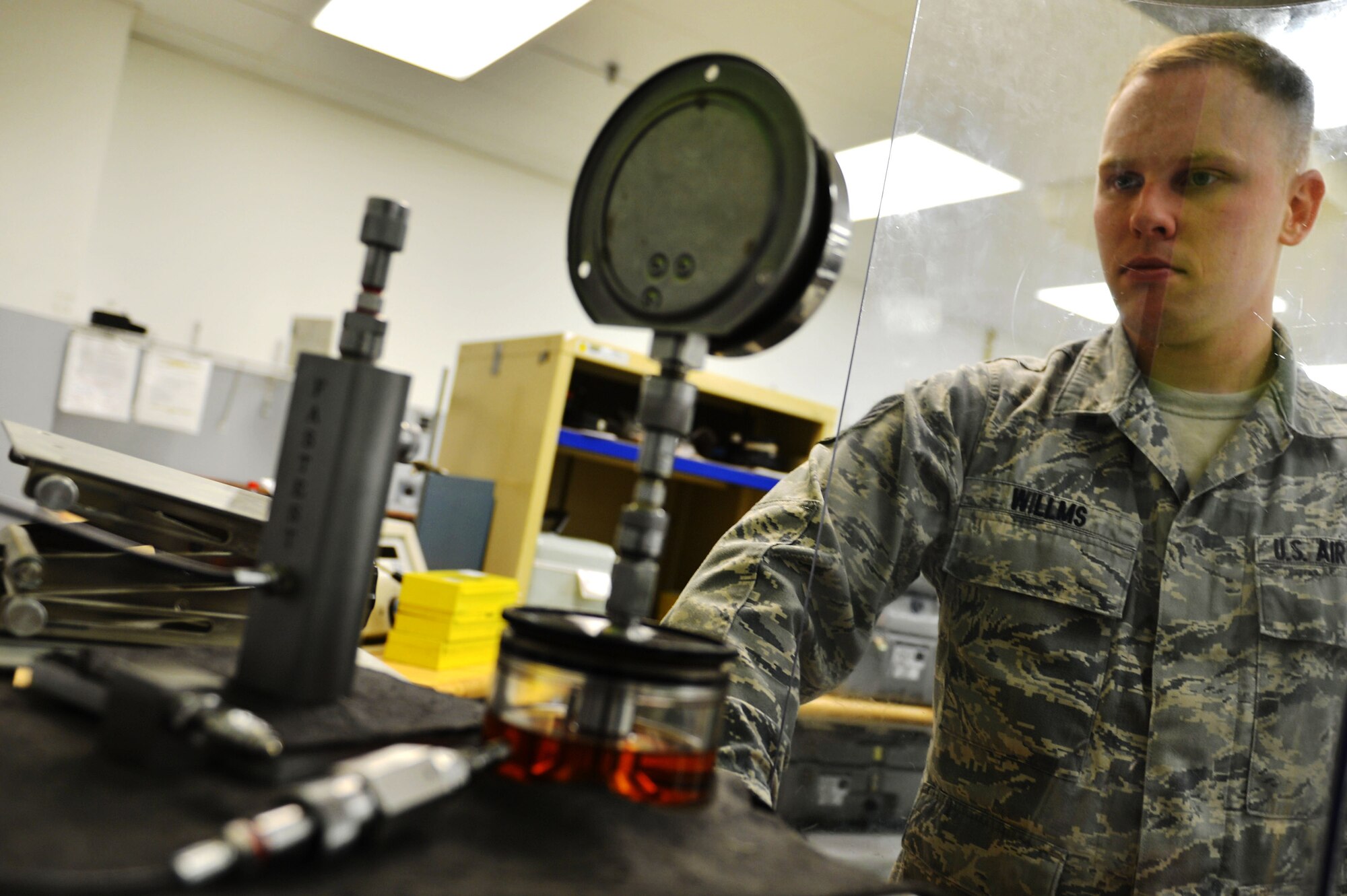 U.S. Air Force Staff Sgt. Zane Wilms precision measurement equipment laboratory technician, calibrates a jack pressure gauge behind a shield at Shaw Air Force Base, S.C., July 11, 2016. The jack pressure gauge is calibrated by PMEL Airmen to allow elevation of an aircraft without the use of its wheels. (U.S. Air Force photo by Airman 1st Class Christopher Maldonado)