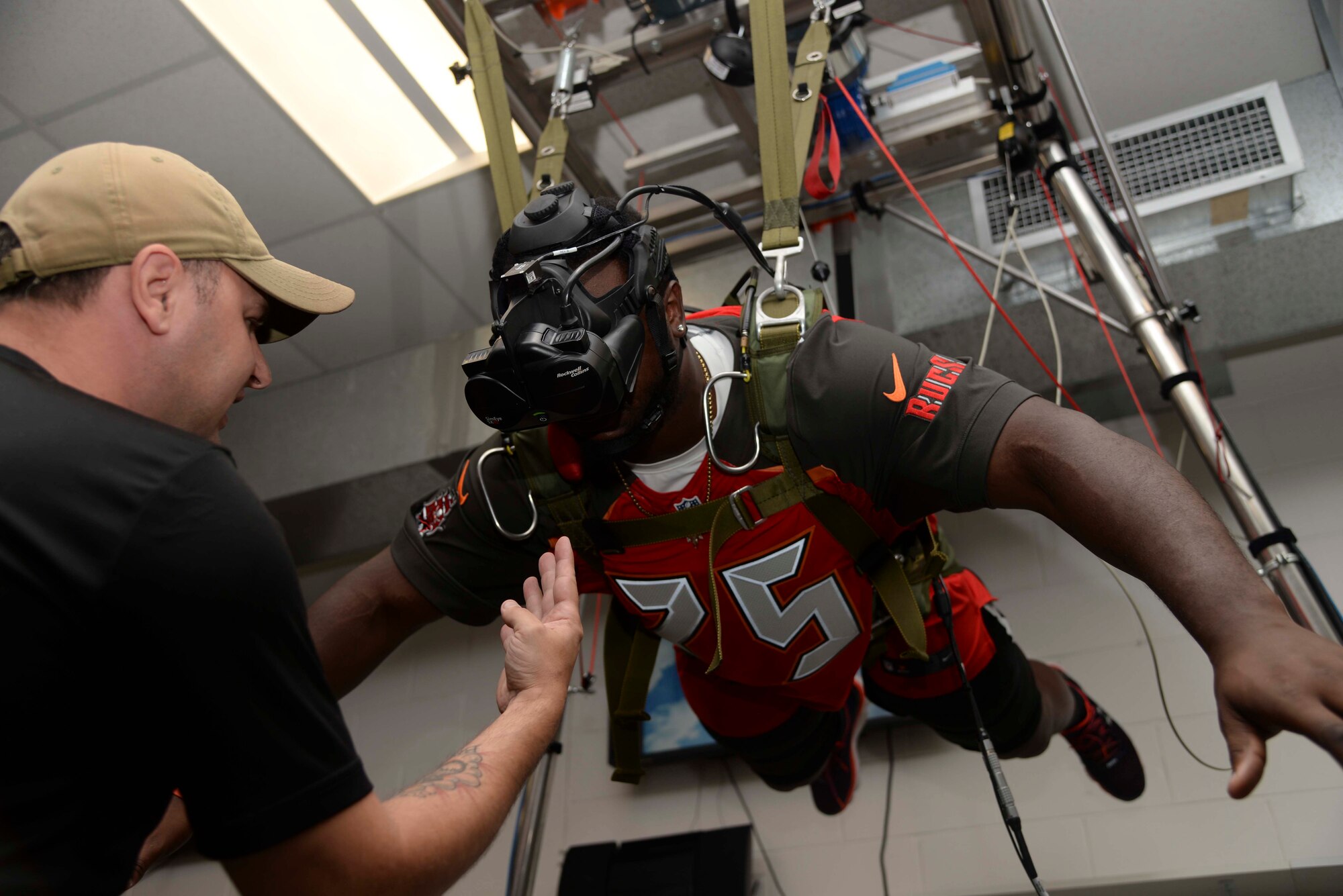 (Left) Sgt. 1st Class Jared Weems, parachute riggers NCO in charge at Special Operations Command Central, assists a rookie from the Tampa Bay Buccaneers on a parachute simulator at MacDill Air Force Base, Fla. June 10, 2016. Some of the rookies tested their airborne skills with virtual reality goggles, which simulated a real jump and landing. 