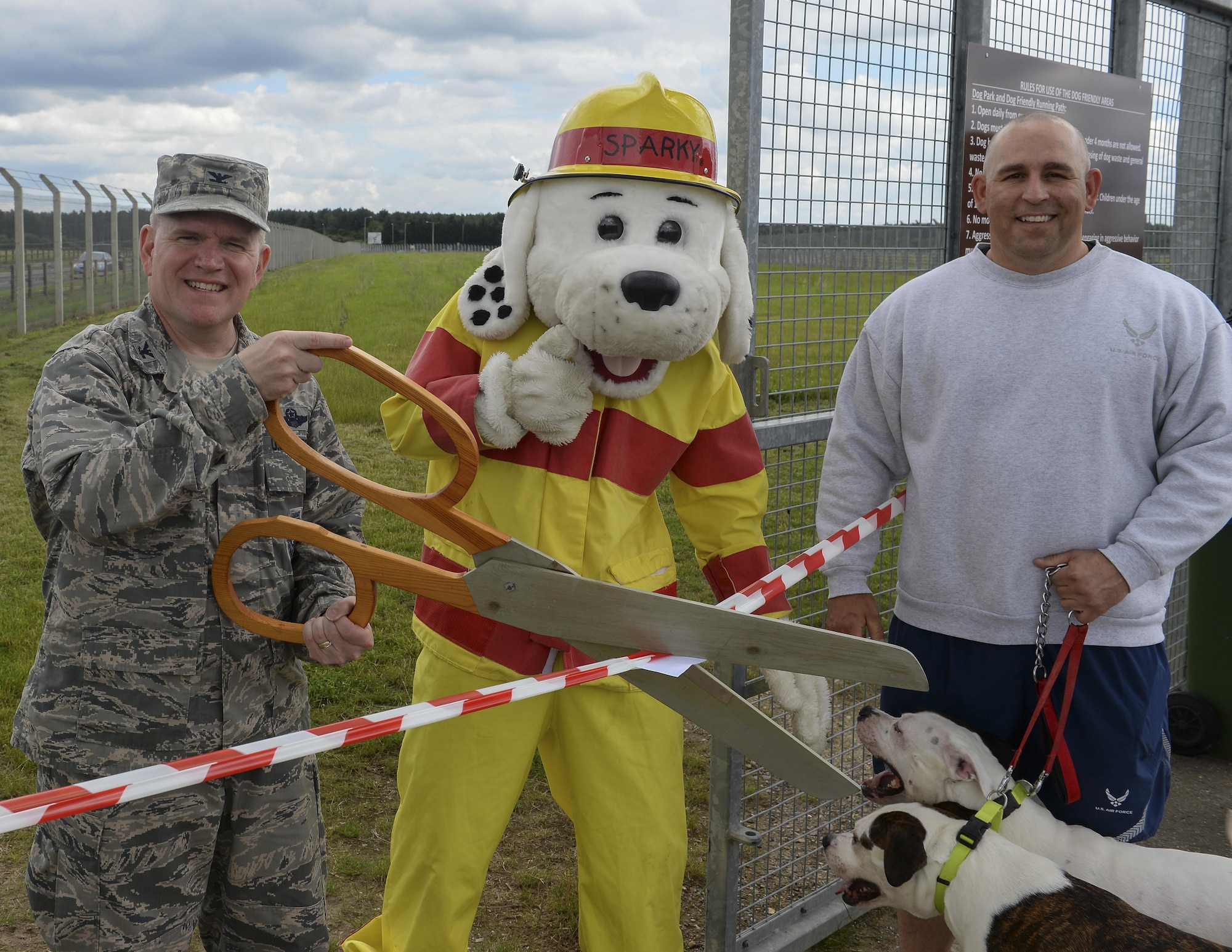 U.S. Air Force Col. Thomas Torkelson, left, 100th Air Refueling Wing commander, cuts the ribbon for the new dog park with Sparky the Fire Dog and Master Sgt. Matthew Deel, 100th Civil Engineer Squadron facility superintendent, June 16, 2016,on RAF Mildenhall, England. The park is open to all members with base access from sunrise to sunset. (U.S. Air Force photo by Staff Sgt. Micaiah Anthony/Released)