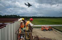 A Charleston C-17 Globemaster III approaches to land June 27 while  Staff Sgt. Martin Lilland, 556th RED HORSE at Hurlburt Field, Florida, and Senior Airman Darrien Anderson, 560th RED HORSE Squadron Joint Base Charleston, South Carolina, position their lift to hang insulation and panels on a 4,800 square-foot preengineered  building building to store RED HORSE construction equipment and gear.  Rapid Engineer Deployable, Heavy Operational Repair Squadron, Engineer (RED HORSE) squadrons provide the Air Force with a highly mobile civil engineering capability in support of contingency and special operations worldwide. They are self-sufficient, mobile squadrons that provide heavy construction support such as runway/facility construction, electrical upgrades, and equipment transport when requirements exceed normal base civil engineer capabilities and where Army engineer support is not readily available. (U.S. Air Force Photo by Michael Dukes)