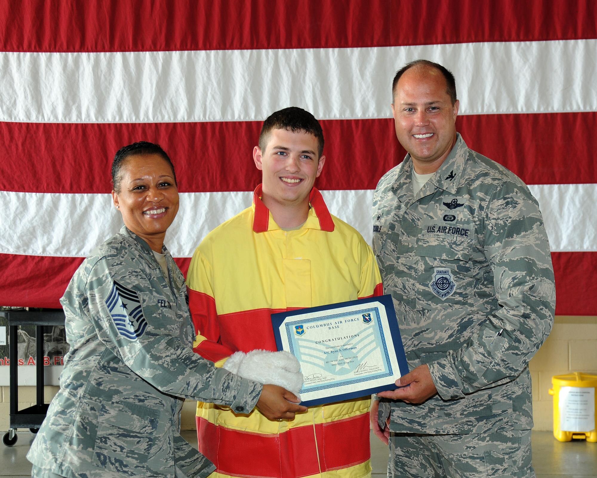 Chief Master Sgt. Rita Felton, 14th Flying Training Wing Command Chief, left, and Col. John Nichols, 14th FTW Commander, left, hand Airman 1st Class Ryan Offerman, 14th Civil Engineer Squadron Firefighter, his Below the Zone certificate June 27 at Columbus Air Force Base, Mississippi. BTZ is the competitive promotion program offered to enlisted personnel in the grade of Airman First Class earning them a promotion six months earlier than normal. Offerman was told to get dressed as the mascot Sparky for children touring the Fire Station but upon his return he was surprised with his notification of BTZ from base leadership and his team. (U.S. Air Force photo/Sharon Ybarra)