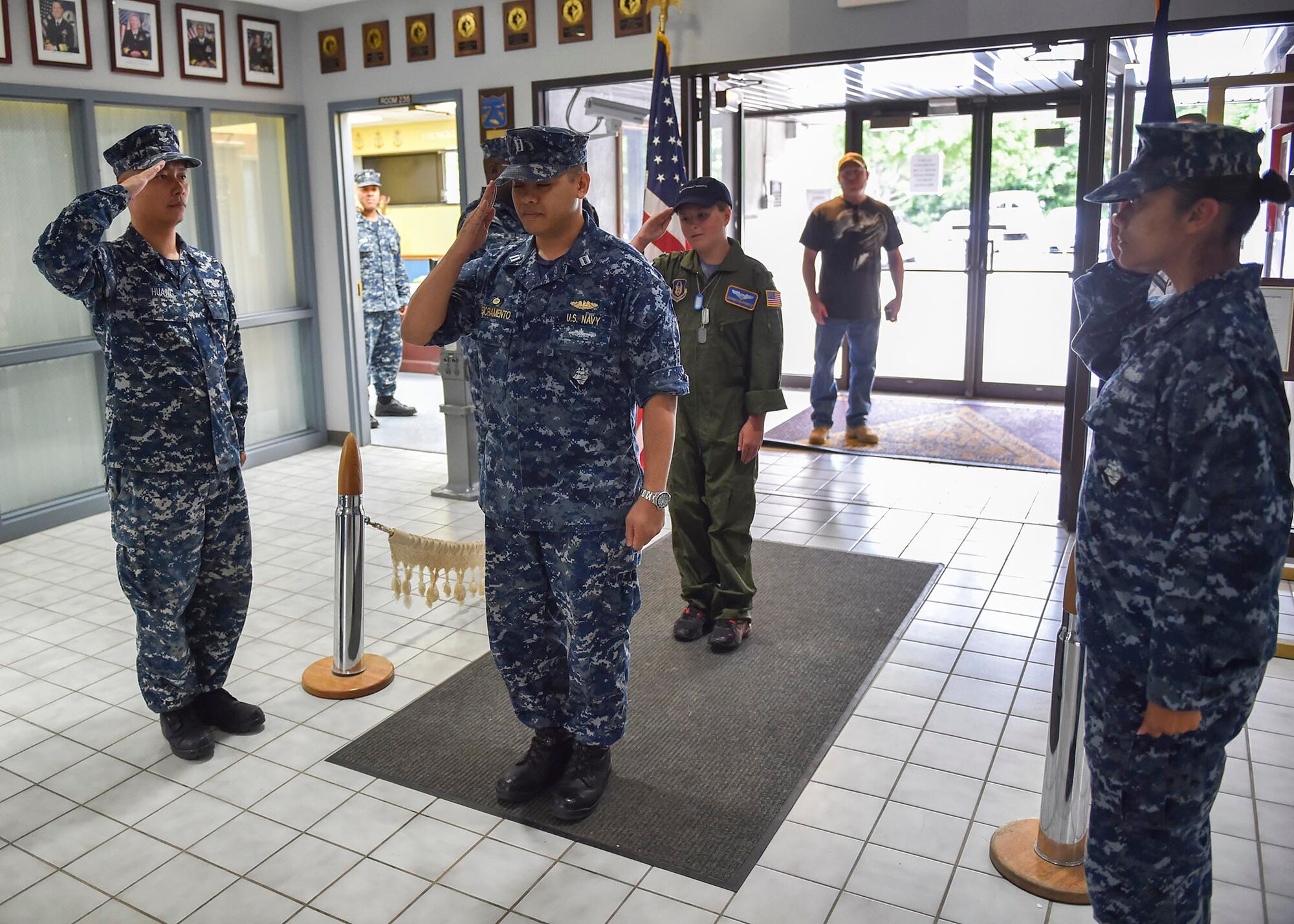 U.S. Navy Lt. Benjamin Sacramento, commanding officer of the Navy Operational Support Center (NOSC) Youngstown, salutes while leading U.S. Air Force Reserve honorary 2nd Lt. Corbin Burggraf, the 910th Airlift Wing’s 63rd Pilot for a Day, in a piping aboard ceremony. Burggraf’s father George watches from the entryway. For Burggraf’s piping aboard, a member of the Navy staff rang a large bell twice, and another member sounded the pipe. The ceremony comes from a Navy tradition of sounding a bell or pipe to announce the boarding of an officer or dignitary. The Pilot for a Day program welcomes children with life threatening or chronic illnesses to Youngstown Air Reserve Station for a day of fun military experiences. (U.S. Air Force photo/Eric White)