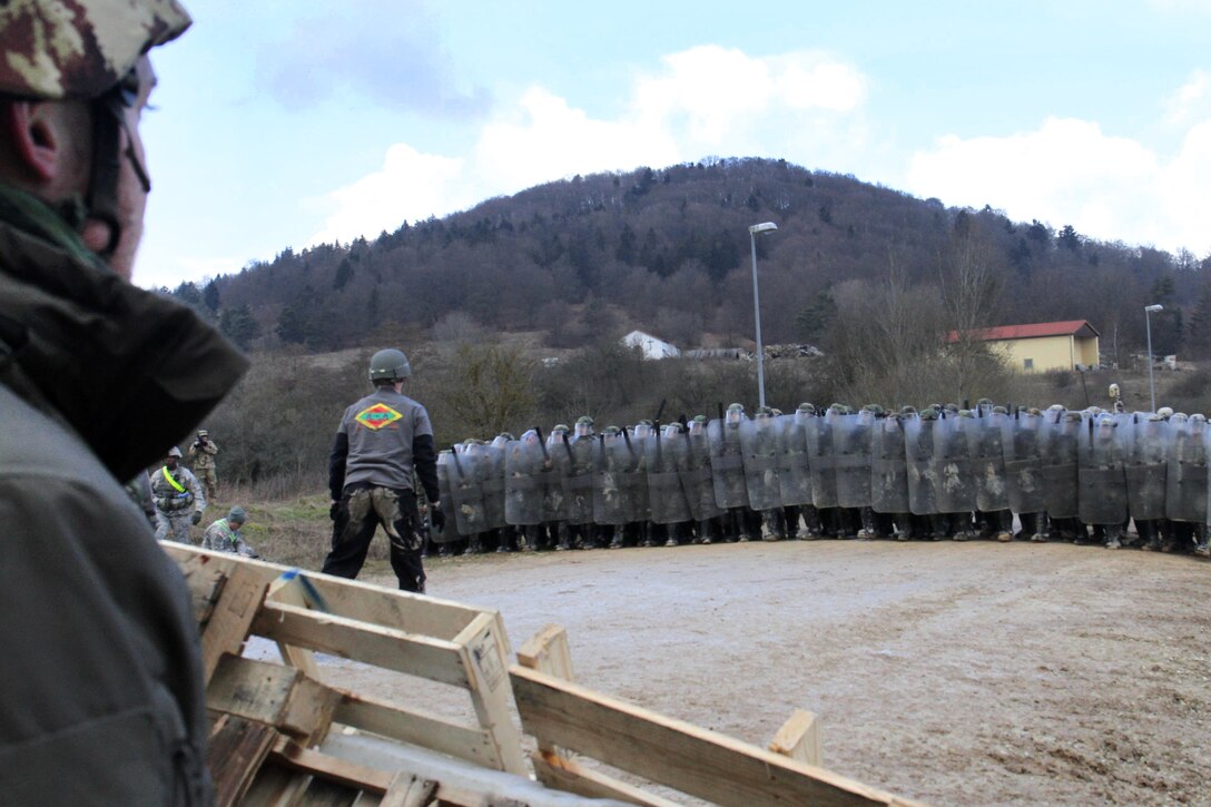 Mock protesters watch as a group of U.S. and Albanian soldiers approach during crowd control training at the Joint Multinational Readiness Center in Hohenfels, Germany, Feb. 25, 2016. Army photo by Staff Sgt. Thomas Duval