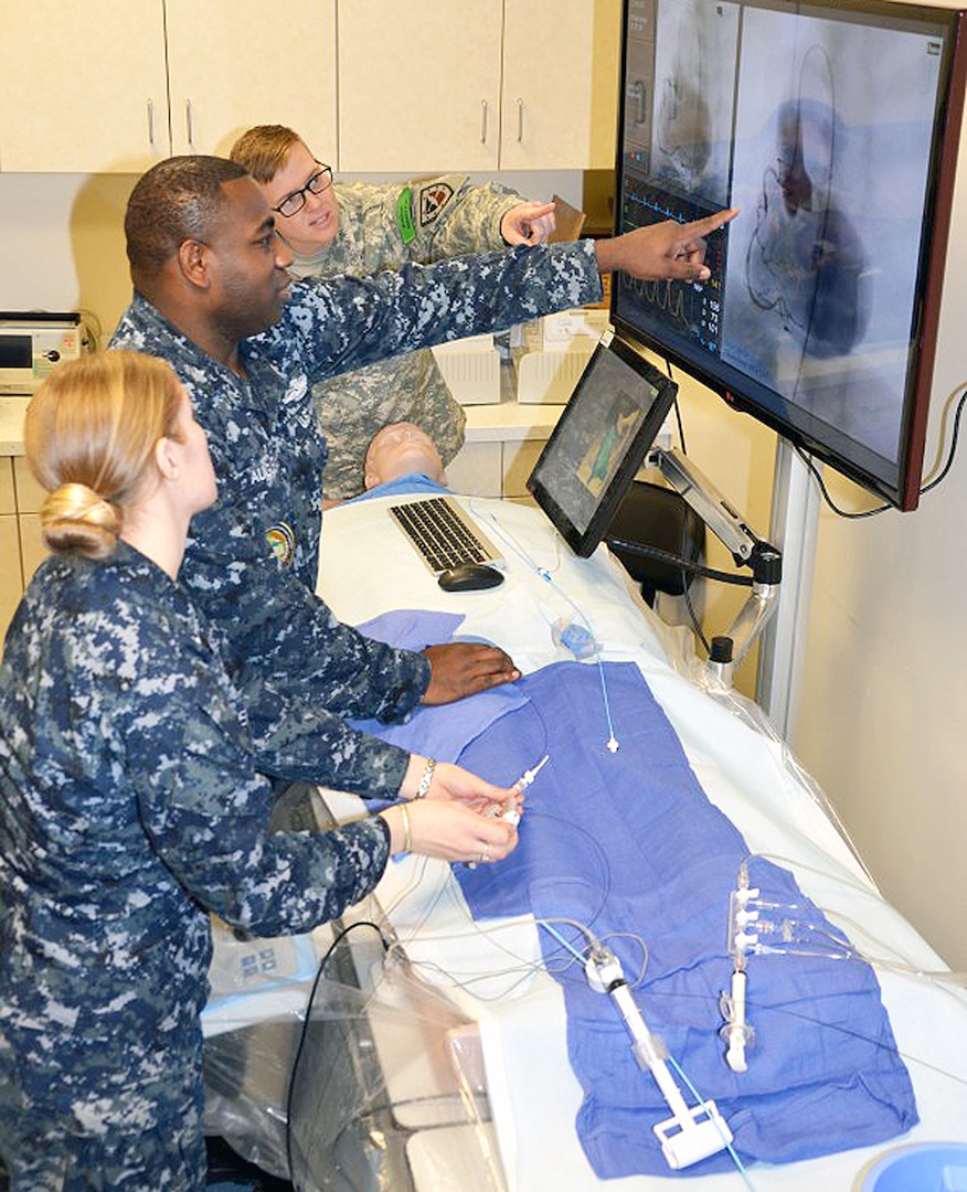 Petty Officer 1st Class Aldrin Augustus (center), cardiovascular technician program instructor and Navy service lead at the Medical Education and Training Campus on Joint Base San Antonio-Fort Sam Houston, instructs Navy Petty Officer 3rd Class Chessa Sheppard (left) and Army Spc. Victoria Belbusti (right) on using the cardiac catheterization simulator during a portion of their CVT didactic training. The simulator is used to demonstrate concepts learned in the classroom and exposes students to hands on training that they will be expected to perform.