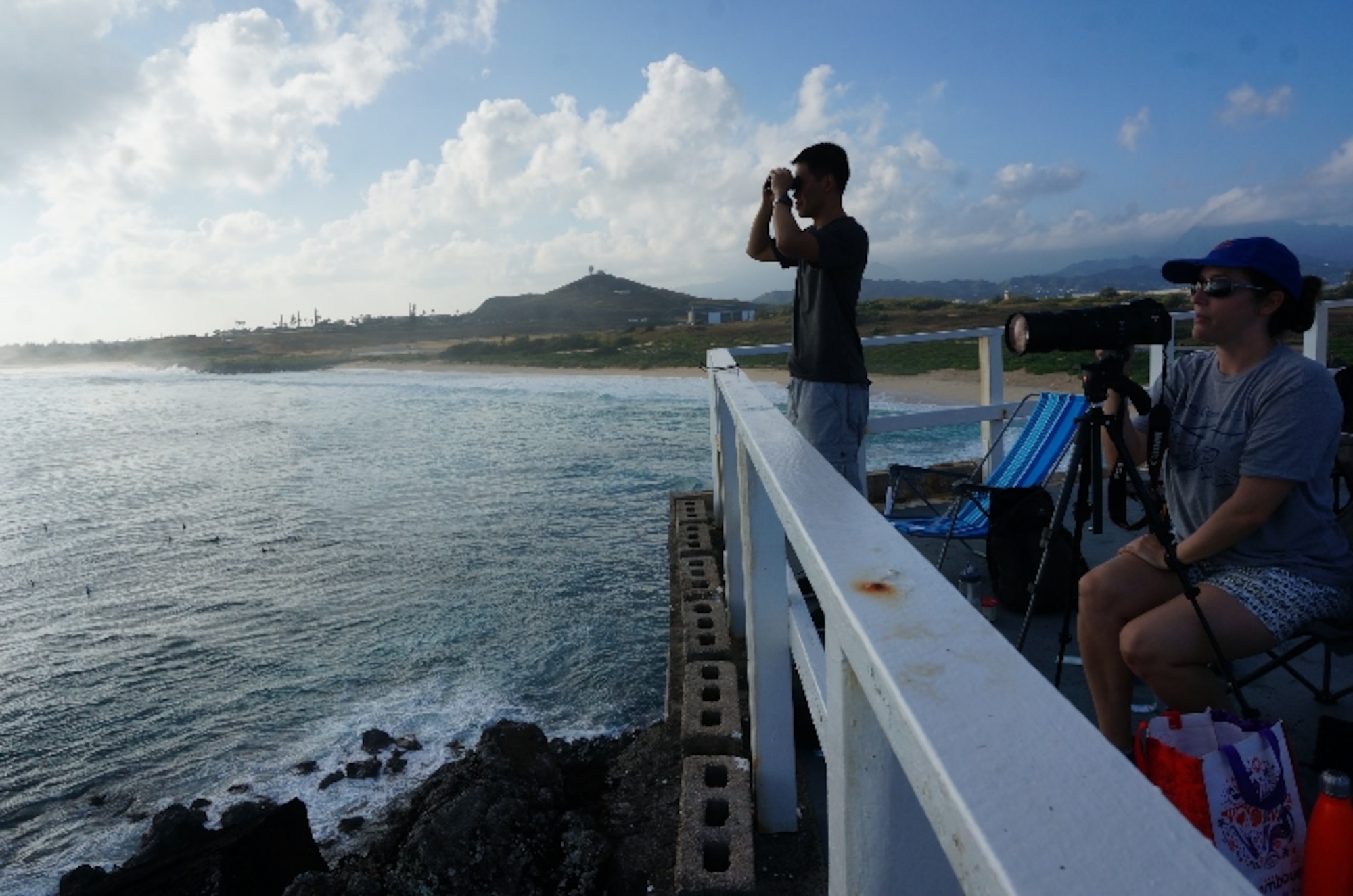 Capt. Aries Guintu, whale count volunteer (left), pans the horizon for humpback whales from atop Marine Corps Base Hawaii's Pyramid Rock Feb. 27, 2016. Guintu is the Communications Division chief for the Air Force Installation Management Support Center's Detachment 2. He joined Sanctuary Ocean Count Site Leader, Capt. Natalie Chounet (seated), for the early-morning venture as surfers pepper the waves below. The Sanctuary Ocean Count project is sponsored by the National Oceanic and Atmospheric Administration to provide scientists with data documenting the humpback whales’ annual 2,500-mile, round-trip migration from Alaska to the Hawaiian Islands. AFIMSC Det. 2 operates out of Joint Base Pearl Harbor-Hickam, Hawaii.  (U.S. Air Force Photo by Senior Master Sgt. Denise Johnson/Released)