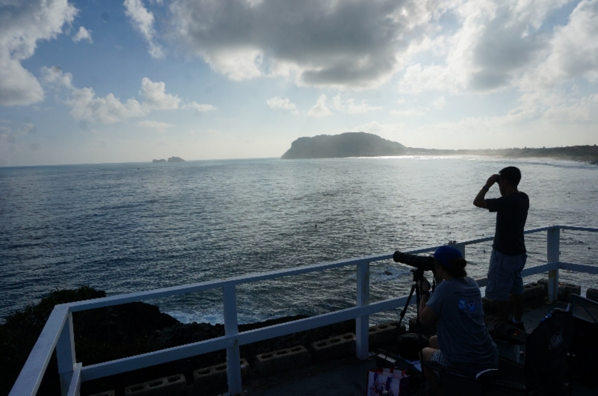 Capt. Aries Guintu, whale count volunteer (right), capitalizes on calm waters and a great vantage point to search for humpback whales from atop Marine Corps Base Hawaii's Pyramid Rock Feb. 27, 2016. Guintu is the Communications Division chief for the Air Force Installation Management Support Center's Detachment 2. He joined Sanctuary Ocean Count Site Leader, Capt. Natalie Chounet (seated), for the early-morning venture as surfers pepper the ocean below. The Sanctuary Ocean Count project is sponsored by the National Oceanic & Atmospheric Administration to provide scientists with data documenting the humpback whales’ annual 2,500-mile, round-trip migration from Alaska to the Hawaiian Islands. Pyramid Rock is off-limits to beachgoers and requires special permission and safety briefings prior to using the facility. (U.S. Air Force Photo by Senior Master Sgt. Denise Johnson//Released)