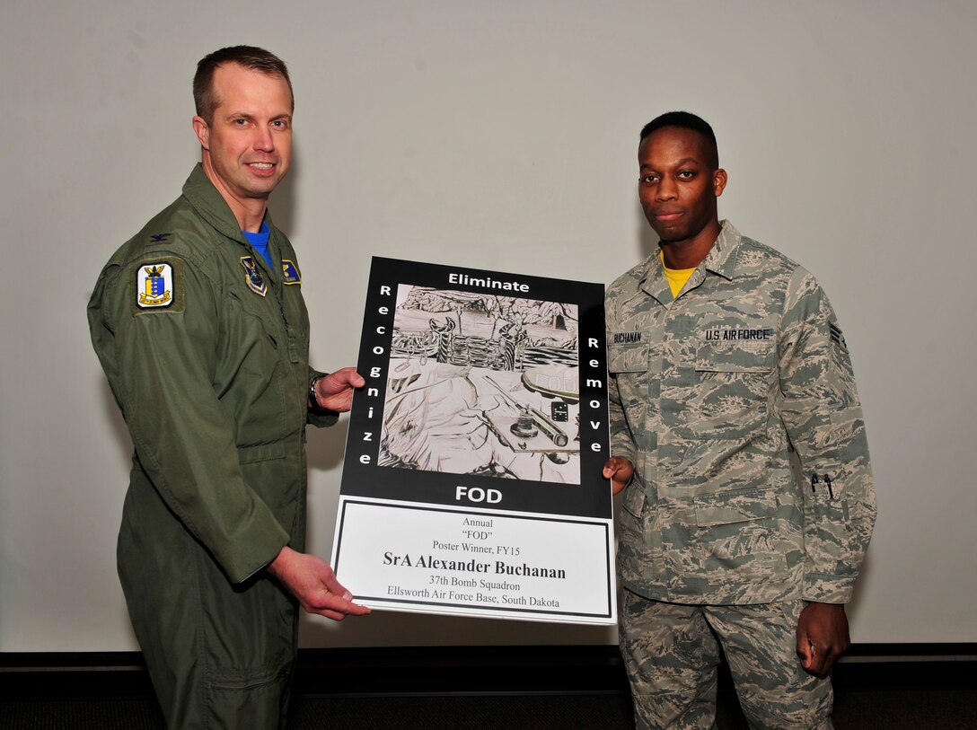 Col. Ty Neuman, 28th Bomb Wing vice commander, left, presents a coin and poster to Senior Airman Alexander Buchanan, 37th Bomb Squadron aviation resource management journeyman,  during an awards presentation at Ellsworth Air Force Base, S.D., March 4, 2016. Buchanan’s poster design won the annual Foreign Object Detection poster contest for fiscal year 2015. The inspiration for the design was described as a perspective change to show how the smallest of foreign objects could cost the Air Force millions of dollars, so these objects were portrayed as big as they appear to ants. (U.S. Air Force photo by Airman 1st Class James L. Miller/Released) 
