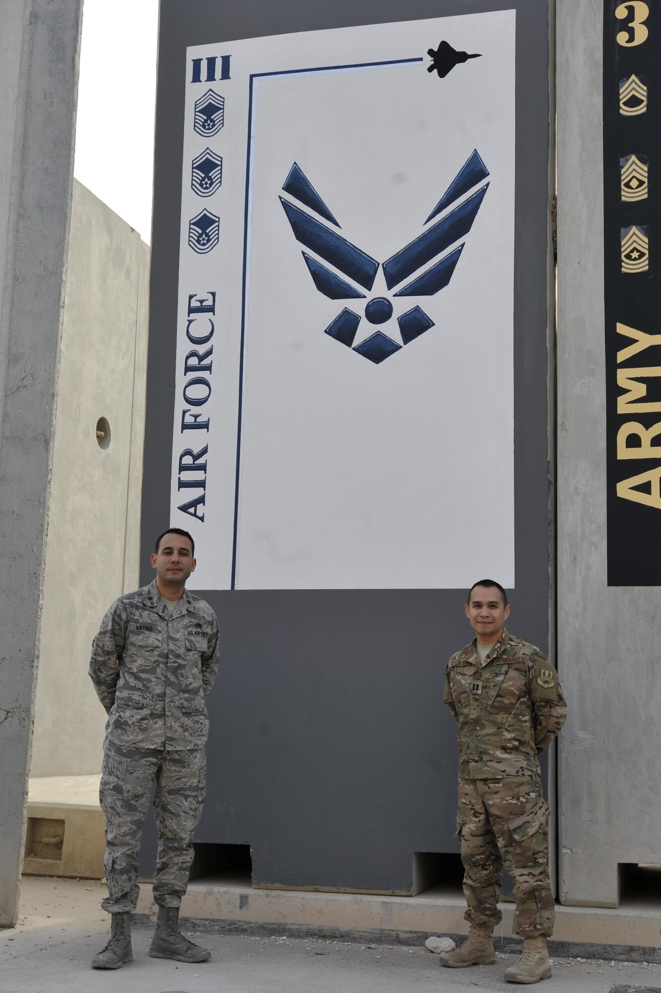 Maj. Juan Matias, Air Forces Central Command's Lessons Learned director, and Capt. Christopher De La Pena, AFCENT's Lessons Learned assistant director, pose for a photograph near a painted Air Force mural March 3 at Al Udeid Air Base, Qatar. The duo assist units in the AFCENT area of operations in preparing, executing and properly documenting operational observations and after action reports to ensure smoother future operations. (U.S. Air Force photo by Spc. Travis Terreo)  