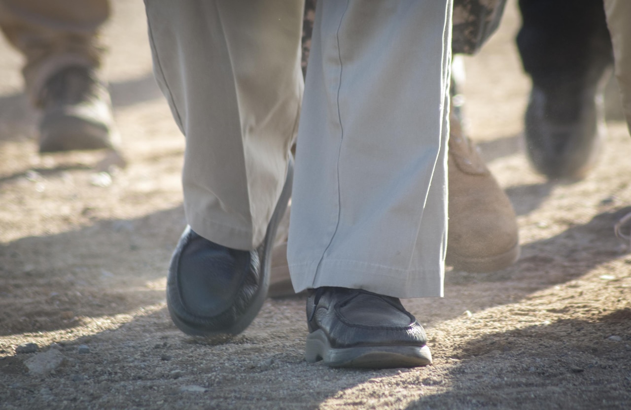 Shoes and slacks of a veteran and several others are shown close up while walking along a dirt path.