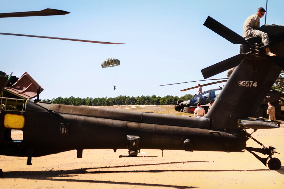 An Air Force combat control school trainee descends during an airborne operation while soldiers reconnect the rotary blades to get an AH-64 Apache helicopter ready for flight at Camp Mackall, N.C., March 17, 2016. Army photo by Capt. Adan Cazarez