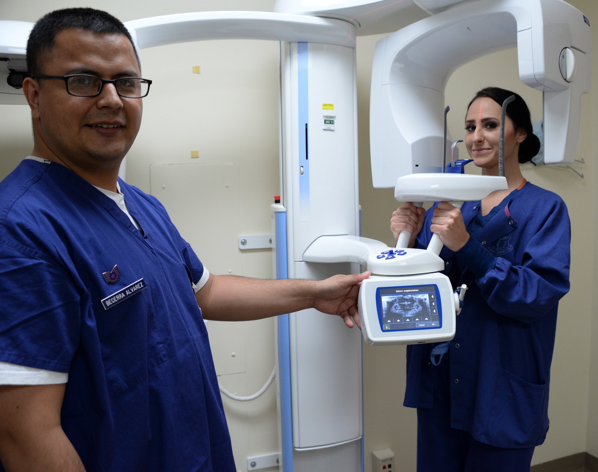 Airman 1st Class Alexandria Stewart, right, with Staff Sgt. Froylan Becerra Alvarez, left, 60th Dental Squadron dental assistants, pose for a photo May 2 at the Arthur B. Sachsel Dental Clinic. Stewart positions herself in the panoramic x-ray machine, which will provide information about missing teeth, additional teeth, sinus issues and jaw joints, to make sure the patient is receiving the best care possible. (U.S. Air Force photo by Senior Airman Amber Carter)