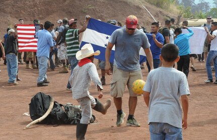 U.S. Air Force Capt. Jesus Lopez, Joint Task Force-Bravo personnel officer, plays soccer with children from Montaña La Oki, a small village located in the mountains east of Comayagua, Honduras, while local village men hold U.S. and Honduran flags welcoming the participants of Chapel Hike 67 and the 3,000 pounds of food they carried 3.4 miles up the mountain, April 30, 2016. During the visit to Montaña La Oki, U.S. service members had the opportunity to watch local school children perform cultural dances, play soccer and hand out the bags of food to villagers as well as some Hondurans who traveled to the location on horseback from the surrounding mountains. (U.S. Air Force photo by Capt. David Liapis/Released)
