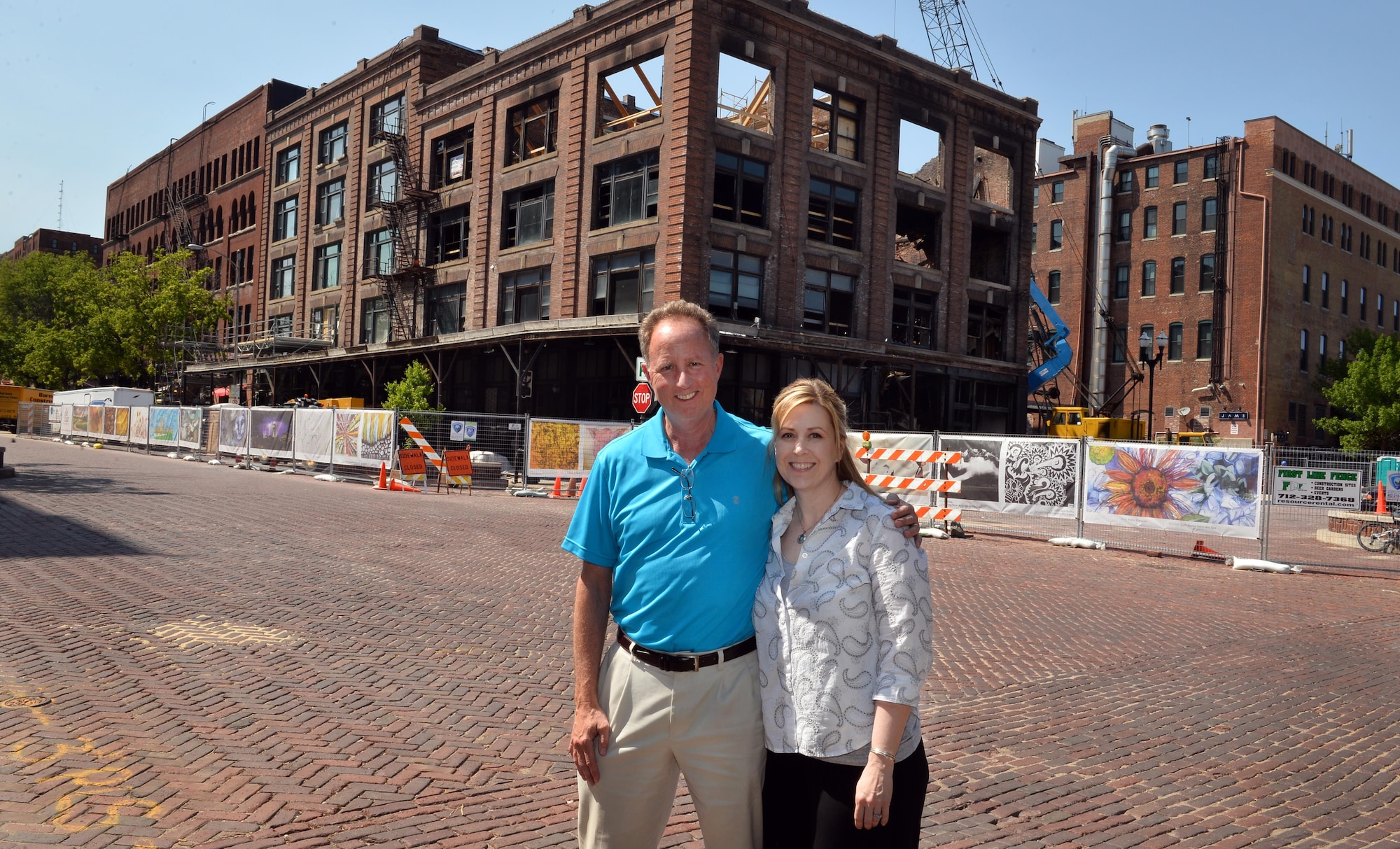 Ken Smith, a weather operations requirements manager with to the 557th Weather Wing, and Lisa Gill, with the 55th Force Support Squadron, stand in front of the Mercer Building in downtown Omaha, Neb., May 5, 2016.  The barrier fence, that surrounds the construction site, is decorated with printed banners depicting Omaha-area artists’ contributions to the Old Market Business Association beautification project designed to shield the site until the restoration is completed.  (U.S. Air Force photo by Josh Plueger/Released)