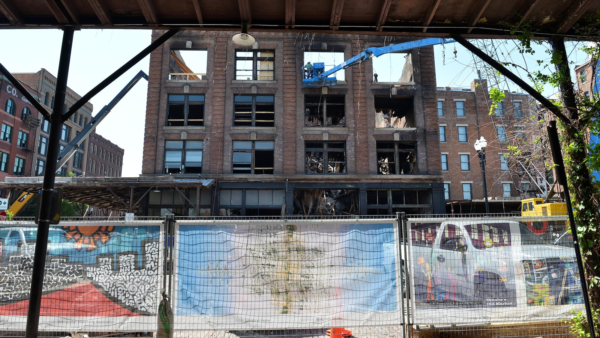Banners, displaying Omaha-area artists’ work, hang along a barrier fence that encircles the renovation efforts of the Old Market Mercer Building in downtown Omaha, Neb., May 5, 2016.  The historic building almost completely burned to the ground from a gas fire in January.  The banners are part of the Old Market Business Association’s beautification project.  (U.S. Air Force photo by Josh Plueger/Released)