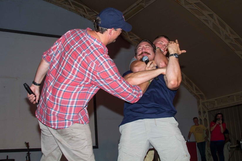 Matthew Lillard (left), actor, holds a microphone for Sgt. 1st Class Timothy Kennedy, a National Guardsman with the 19th Special Forces Group and a mixed martial artist with the Ultimate Fighting Championship, while he instructs Spc. Logan Kelly, a carpenter with 1194th Engineer Company, 682nd Engineer Battalion, on how to properly preform a rear naked choke during the USO May Madness Tour on Camp Arifjan, Kuwait, May 18, 2016. The celebreties on the tour said they wanted to repay Servicemembers for their sacrifice and dedication to America.