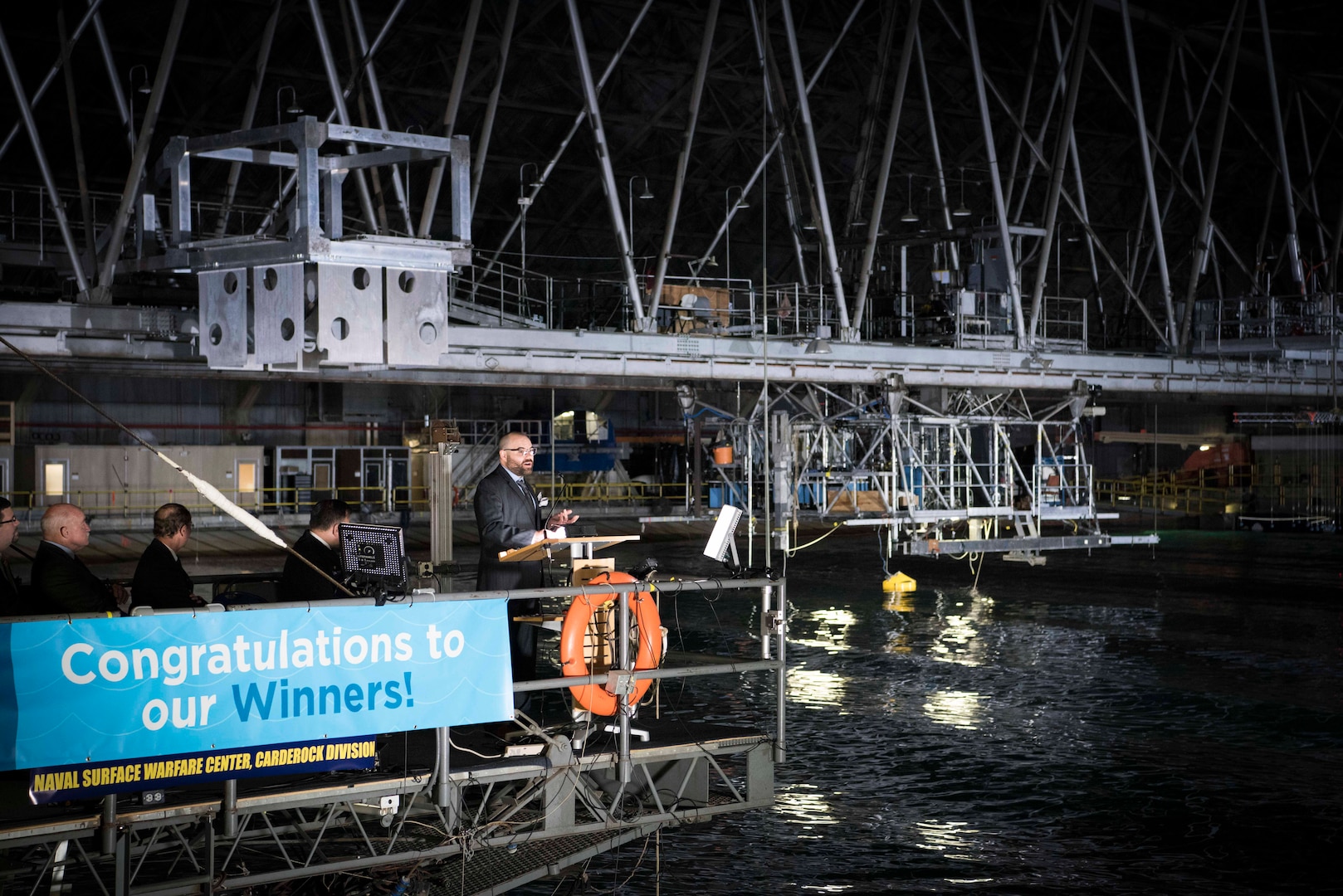 Alex Hagmuller, a mechanical engineer from the Portland, Oregon-based team AquaHarmonics, tells the crowd how their wave energy converter (WEC) is working to absorb energy from waves, which can then be converted and used as a power source. AquaHarmonics competed against eight other teams to win the 2016 Wave Energy Prize of $1.5 million, which was awarded Nov. 16 at Naval Surface Warfare Center, Carderock Division in West Bethesda, Md. The final testing stage took place at the Maneuvering and Seakeeping (MASK) Basin, Carderock's state-of-the-art indoor ocean. The Wave Energy Prize is a public prize competition sponsored by the U.S. Department of Energy (DoE), and supported by the Office of Naval Research (ONR) and Carderock. (U.S. Navy photo by Monica McCoy/Released)