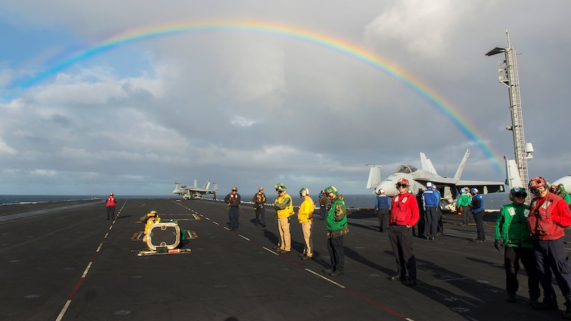 Sailors prepare to conduct operations from the flight deck of  a ship with a rainbow in the background.
