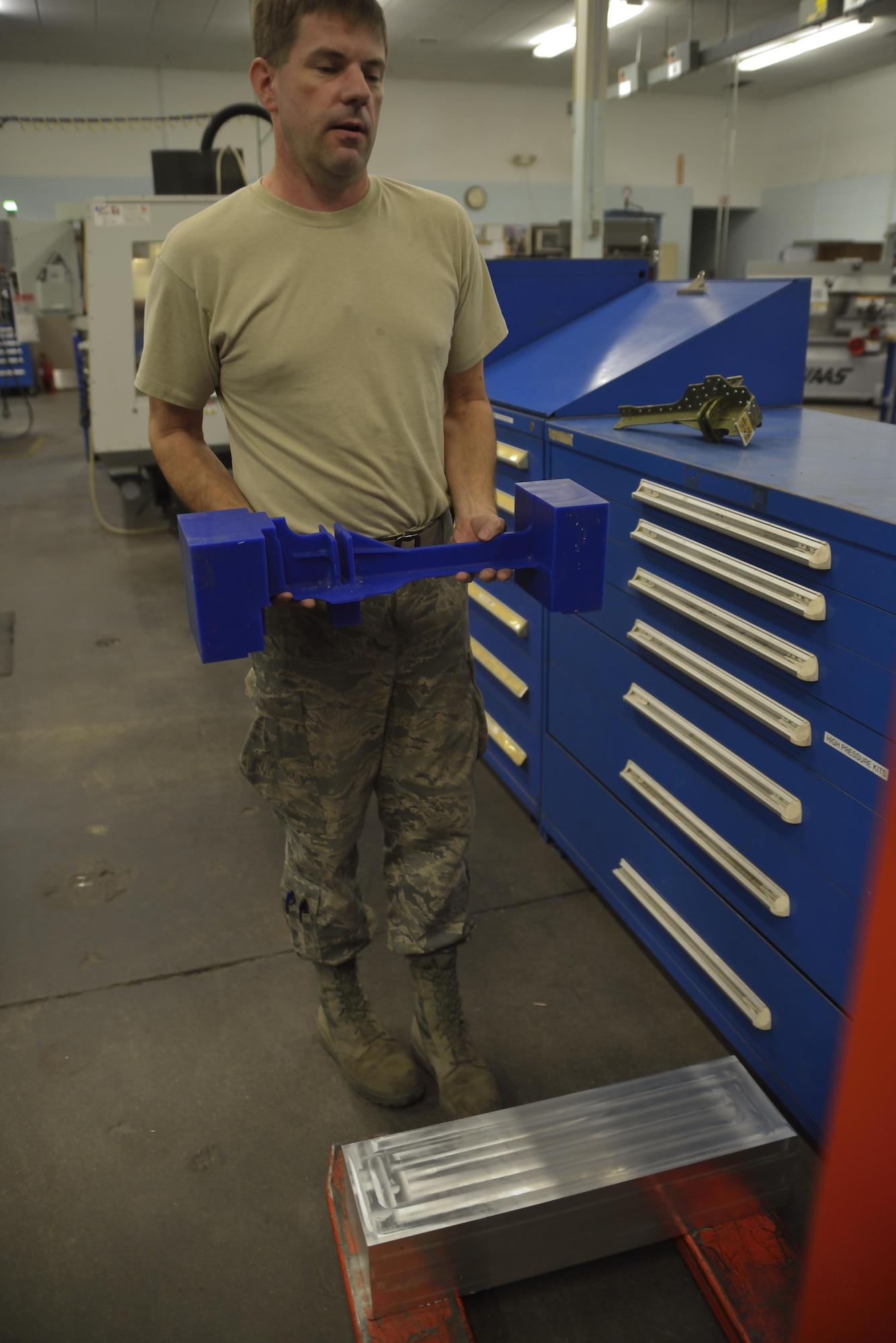 A blue-wax test model of the support bracket is being held by TSgt. Richard Towlson, assistant NCOIC of the Machine shop and at his feet is the 100 lb. block of metal, that is similar to the one used to make the bracket.