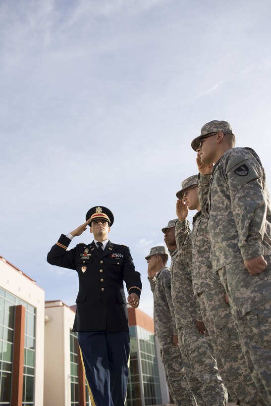 A first lieutenant Officer wearing dress blues saluting soldiers wearing ACU outside during the day.