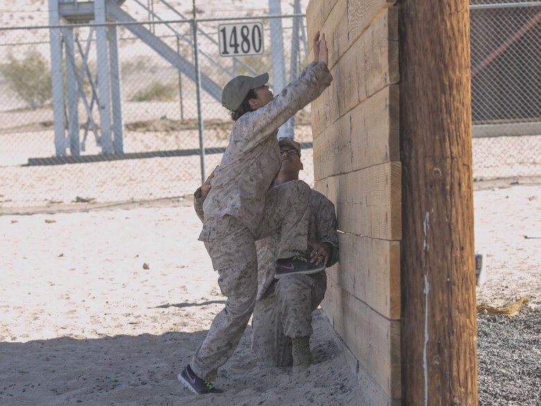 Cpl. Joseph Aguilar, rifleman, 1st Battalion, 7th Marine Regiment, helps his wife, Esther, over the wall at the obstacle course during the battalions Jane Wayne Day aboard Marine Corps Air Ground Combat Center, Twentynine Palms, Calif., Oct. 14, 2016. (Official Marine Corps photo by Cpl. Thomas Mudd/Released)