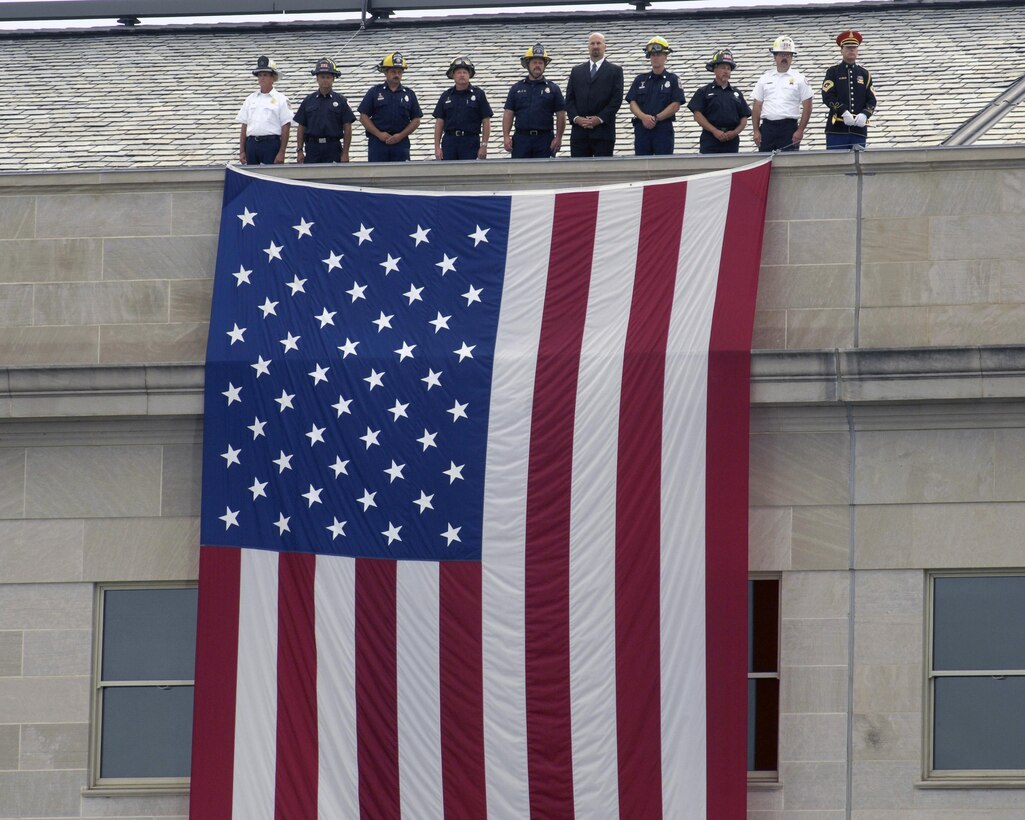 During ceremonies dedicating the Pentagon Memorial, representatives of police and firefighting units who were the first to respond to the 9/11 terrorist attack on the Pentagon, stand at the top of a large American flag suspended from the roof of the Pentagon, Sept. 11, 2008, just as they stood seven years earlier. DoD photo by R. D. Ward 