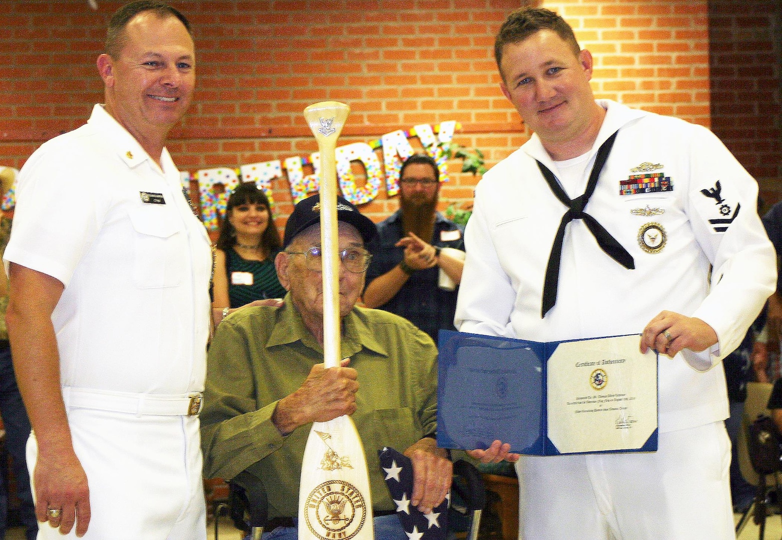Senior Chief Petty Officer Daniel Christmas (left) and Petty Officer 2nd Class Keyth Curnutt (right) of the Navy Recruiting District San Antonio present 100-year-old Navy veteran Tom Littleton with an American flag and a special Navy paddle at a special celebration in Devine.