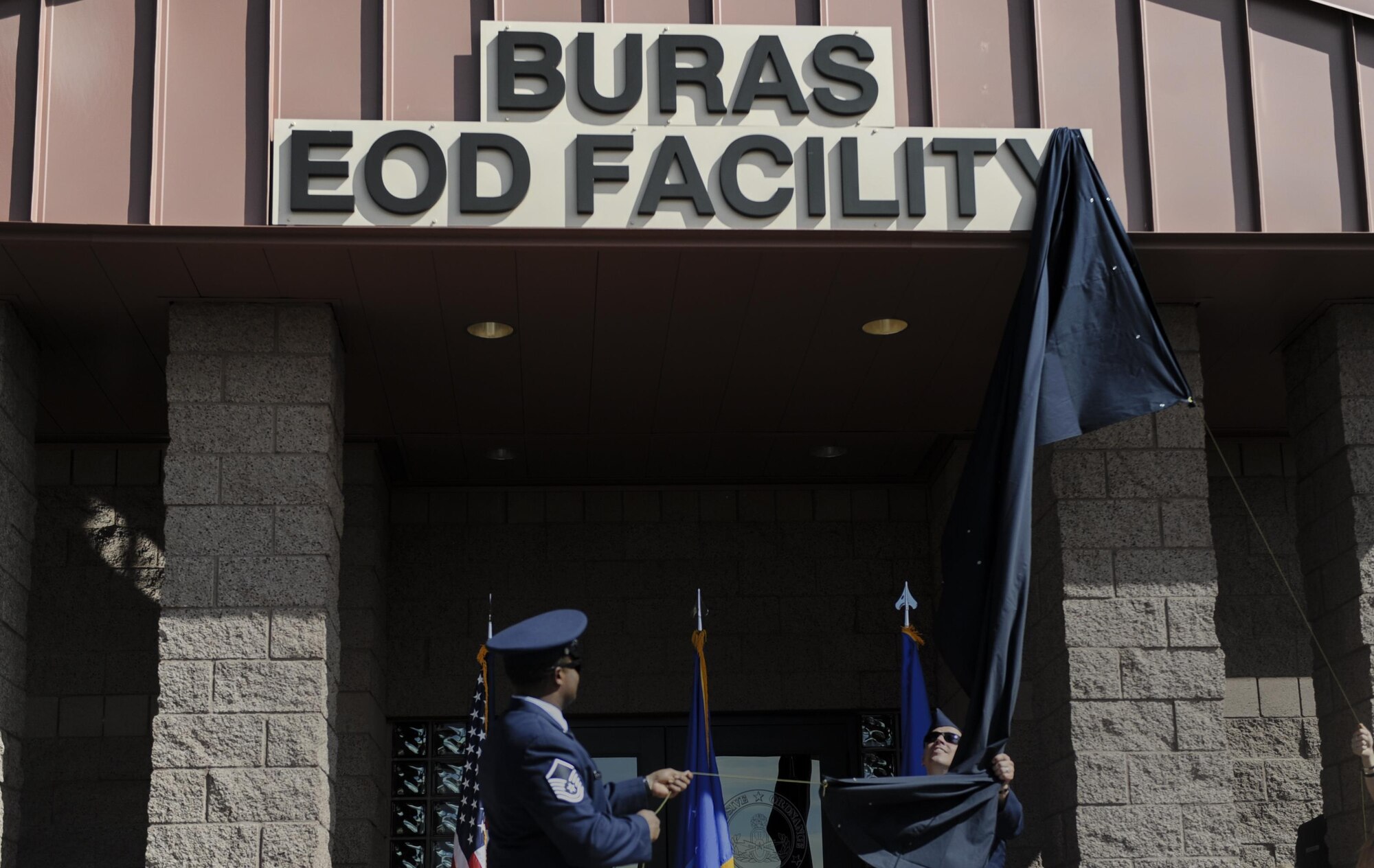 Family of Senior Airman Michael Buras and Explosive Ordinance Disposal technicians pull back a cover to reveal a new EOD building sign at Nellis Air Force Base, Nev., Sept. 21. The 99th Civil Engineer Squadron Explosive Ordinance Flight paid respect to Buras by naming and dedicating their newest facility in his honor on Nellis Air Force Base, Nev., during a ceremony, Sept. 21. (U.S. Air Force photo by Airman 1st Class Kevin Tanenbaum/Released)