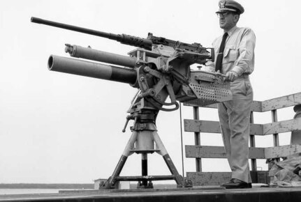 "Chief [Warrant] Gunner Elmer L. HICKS, USCG, has designed an adapter for a .50 caliber Browning machine gun on top of the recoil cylinder of the 81mm seagoing mortar mount which may eventually be standard armament on all Coast Guard cutters.  The accompanying picture shows Chief Gunner HICKS beside his mount.  The 81mm is designated a mortar but it can also be fired by trigger.  It is light weight, has a simple pedestal mount and can be operated in train and elevation by one man.  A variety of ammunition types are available to this gun making it a very effective weapon for WPB Class Coast Guard Cutters.  The stability provided by using the 81mm mortar as a platform also increases the accuracy of the .50 caliber gun."
No date; photographer unknown.