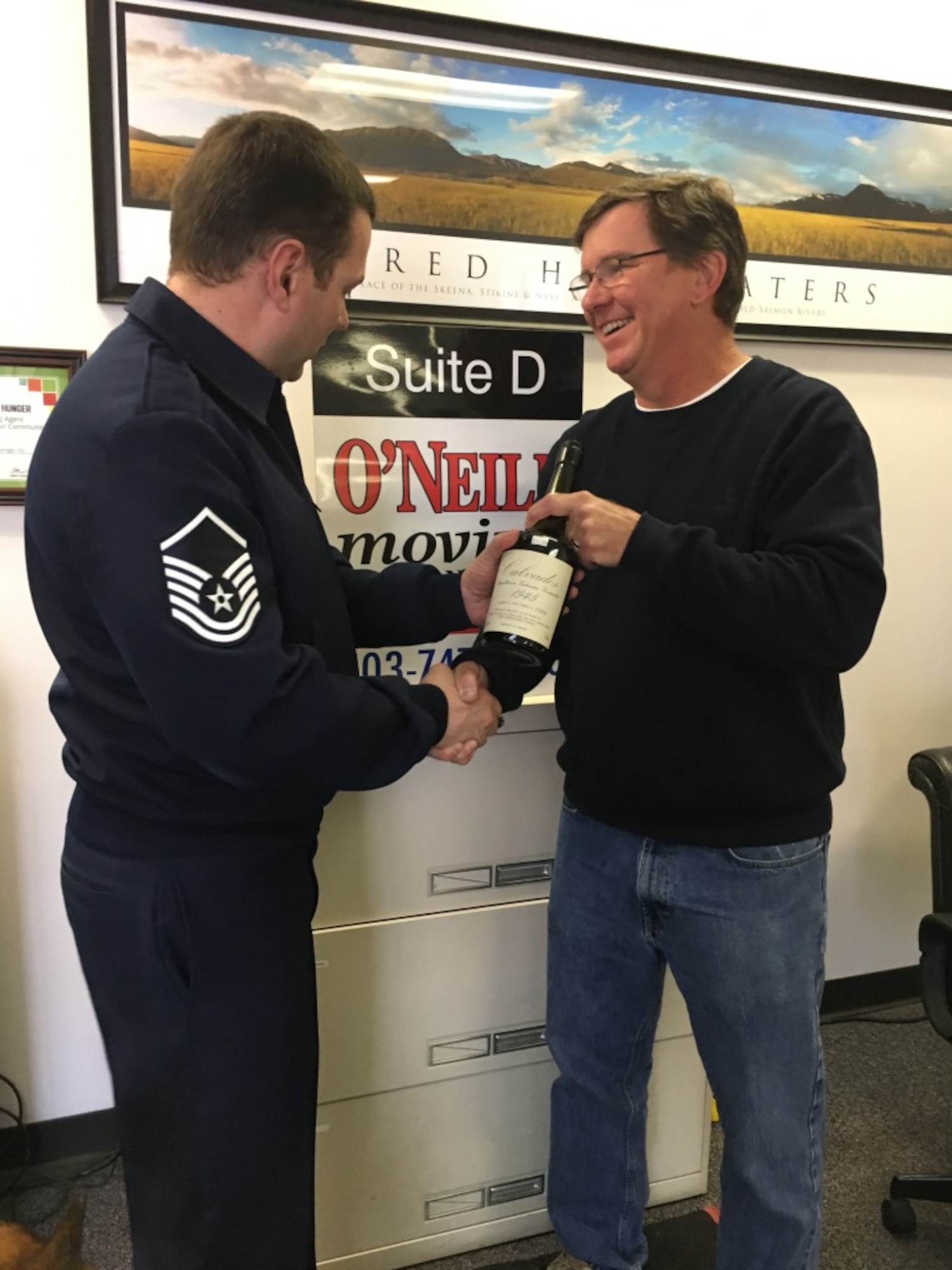 Mr. Brad Boland (right), President O-Neill Transfer and Storage Co., hands off a bottle of 1945 Calvados brandy to Master Sgt. Todd Wivell, 62nd Airlift Wing chief of public affairs, March 5, 2017 at the O-Neill business office in Beaverton, Oregon. Boland, along with other members from the east coast to the west coast, assisted in transporting two separate bottles of this brandy in order to help two World War II veterans complete a pact to toast their fallen comrades. (Courtesy photo
