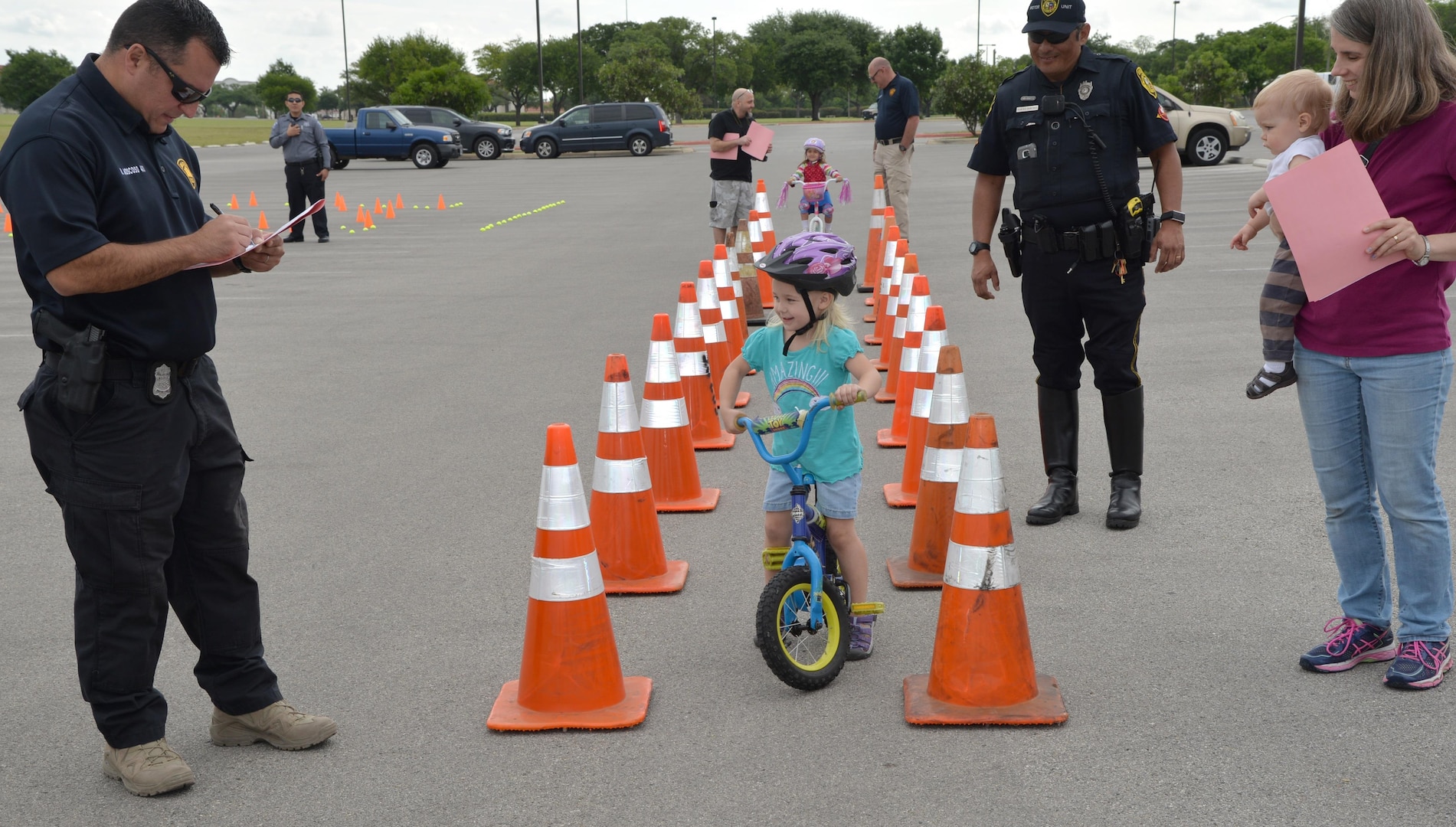 Erika, 3 years old, was the first to start off the obstacle course at the 2017 Spring Bike Rodeo April 8 at Joint Base San Antonio-Fort Sam Houston .  Overlooking her at the finish line was (right) her Mother Carolyn and baby brother, (right back). Officer Ramiro Sanchez, Bexar County Sherriff Traffic Control and (Left) Officer W. Moscoso, San Antonio Police Department tally her score at the finish line.