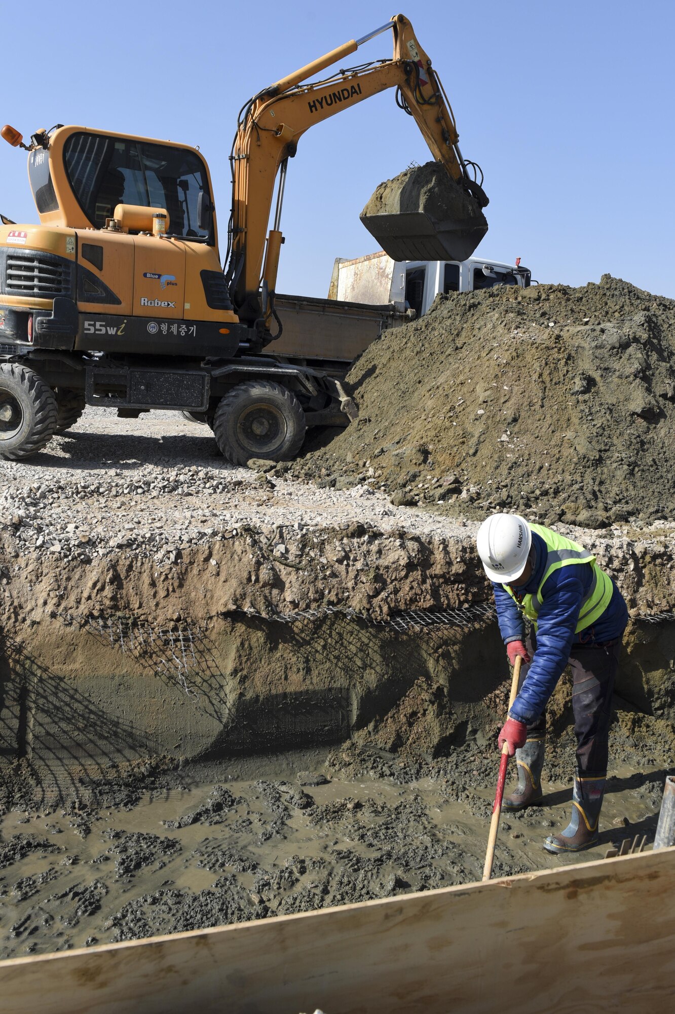 Mun Sung-Hwan, Republic of Korea contractor, rakes the ground while Park Chul-Hee, Republic of Korea contractor, uses an excavator to remove dirt from a trench at Kunsan Air Base, Republic of Korea, March 21, 2017. The trench will house fuel lines that will run to the new flow-through shelters for the 35th Fighter Squadron at Kunsan. (U.S. Air Force photo by Senior Airman Michael Hunsaker/Released)