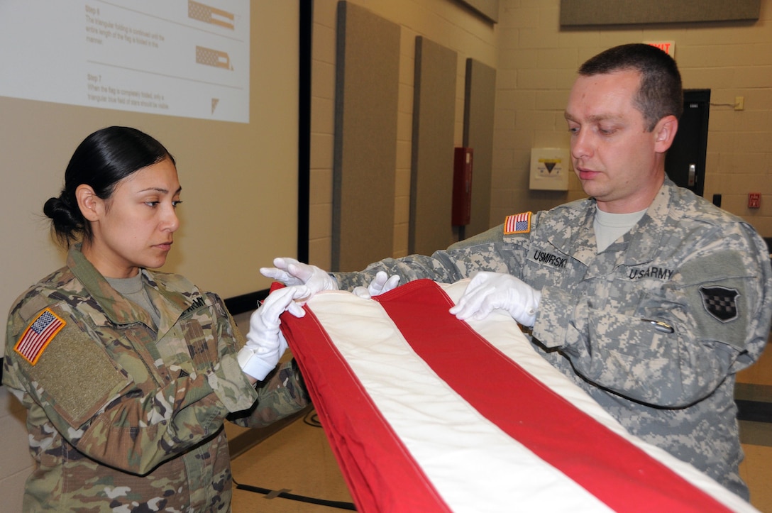 Staff Sgt. Lucia Lopez, a petroleum supply specialist from Orange, New Jersey currently assigned to the U.S. Army Reserve’s 277th Quartermaster Company, receives assistance from Staff Sgt. Alex Ushomirsky learning how to properly fold the U.S. flag April 20 during military funeral honors training at 99th Regional Support Command headquarters on Joint Base McGuire-Dix-Lakehurst, New Jersey.  Ushomirsky is a paralegal currently working in Casualty Operations for the 99th RSC.  The 99th RSC supports the casualty mission by providing support and assistance to families of Soldiers and Department of Army civilians reported as a casualty (deceased, missing, or whereabouts unknown).