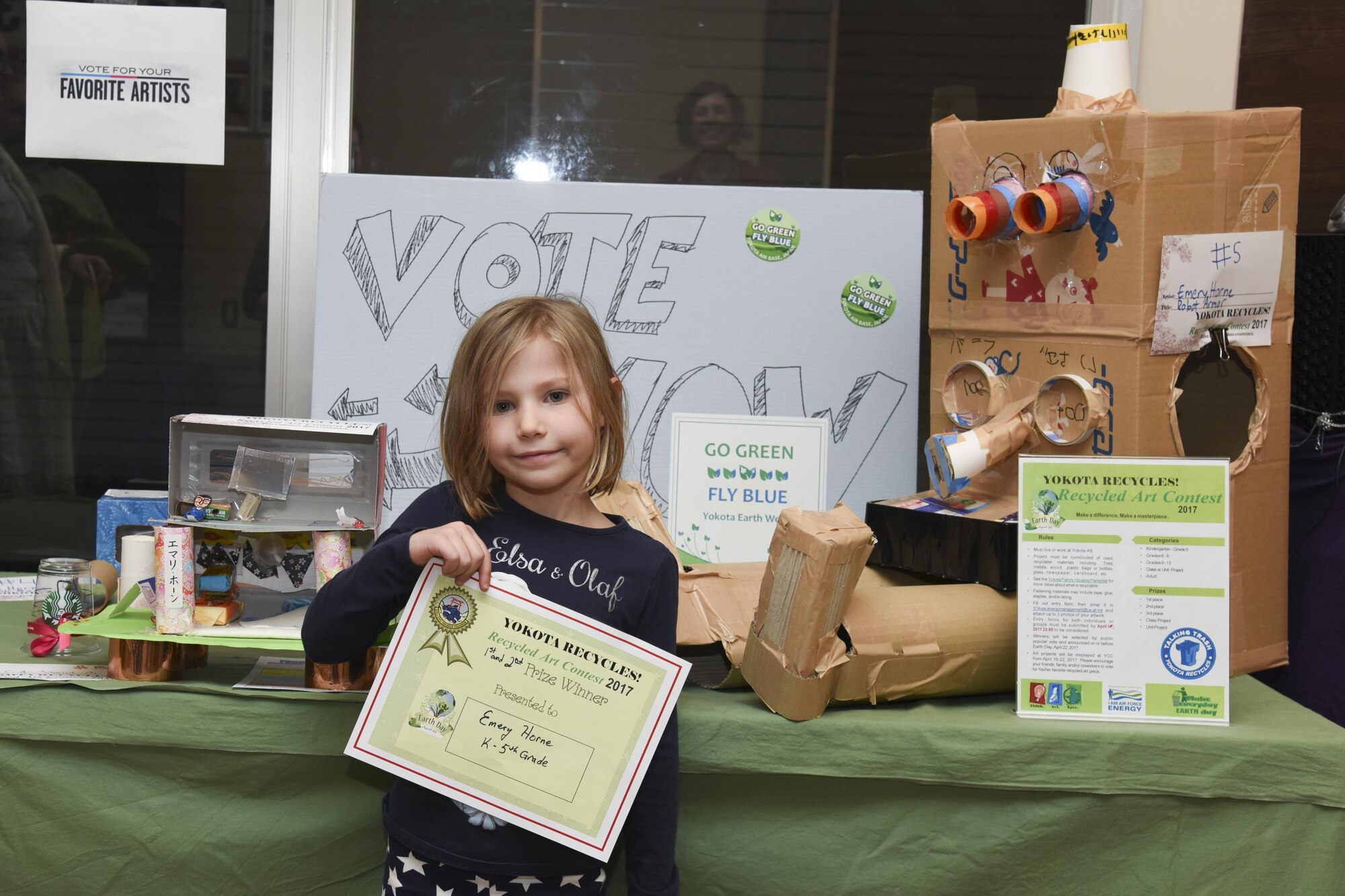 Emery Horne, daughter of Jennifer Horne, poses in front of her works of art after receiving first and second place award certificates in the individual category during the Recycled Art Contest April 22, 2017, at Yokota Air Base, Japan. Horne created a Robot Armor and a Green Dollhouse for the contest. (U.S. Air Force photo by Machiko Arita)