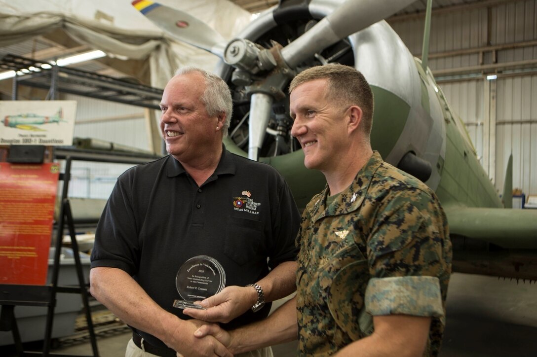 Robert Cramsie, left, a restoration volunteer with the Flying Leatherneck Aviation Museum and a board member of the Flying Leatherneck Historical Foundation, displays his award while shaking hands with Col. Jason Woodworth, commanding officer of Marine Corps Air Station Miramar, Calif., at the Flying Leatherneck Aviation Museum Restoration Facility at MCAS Miramar, Calif., April 21. 
Cramsie received the Northrop Grumman Excellence in Volunteerism Award for volunteering more than 2,500 hours to restore a last-of-its-kind Douglas SBD-1 Dauntless dive bomber, reconstructed using blueprints, salvaged and self-fabricated parts. (U.S. Marine Corps photo by Lance Cpl. Liah Kitchen/Released)