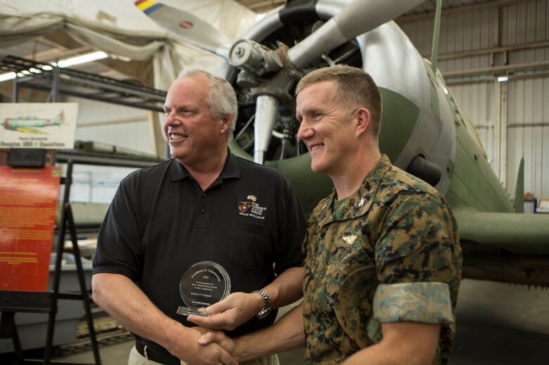 Robert Cramsie, left, a restoration volunteer with the Flying Leatherneck Aviation Museum and a board member of the Flying Leatherneck Historical Foundation, displays his award while shaking hands with Col. Jason Woodworth, commanding officer of Marine Corps Air Station Miramar, Calif., at the Flying Leatherneck Aviation Museum Restoration Facility at MCAS Miramar, Calif., April 21. 
Cramsie received the Northrop Grumman Excellence in Volunteerism Award for volunteering more than 2,500 hours to restore a last-of-its-kind Douglas SBD-1 Dauntless dive bomber, reconstructed using blueprints, salvaged and self-fabricated parts. (U.S. Marine Corps photo by Lance Cpl. Liah Kitchen/Released)