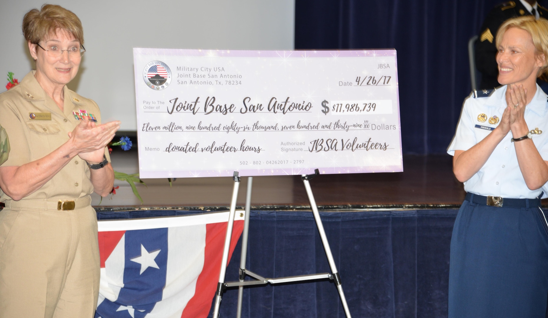 Rear Adm. Rebecca McCormick-Boyle, Navy Medicine Education, Training and Logistics Command commander (left) and Brig. Gen. Heather Pringle, 502nd Air Base Wing and Joint Base San Antonio commander (right) applaud a check showing the monetary value of donated volunteer hours, $11.9 million, from JBSA members in 2016 at the JBSA Volunteer of the Year Awards ceremony April 26.