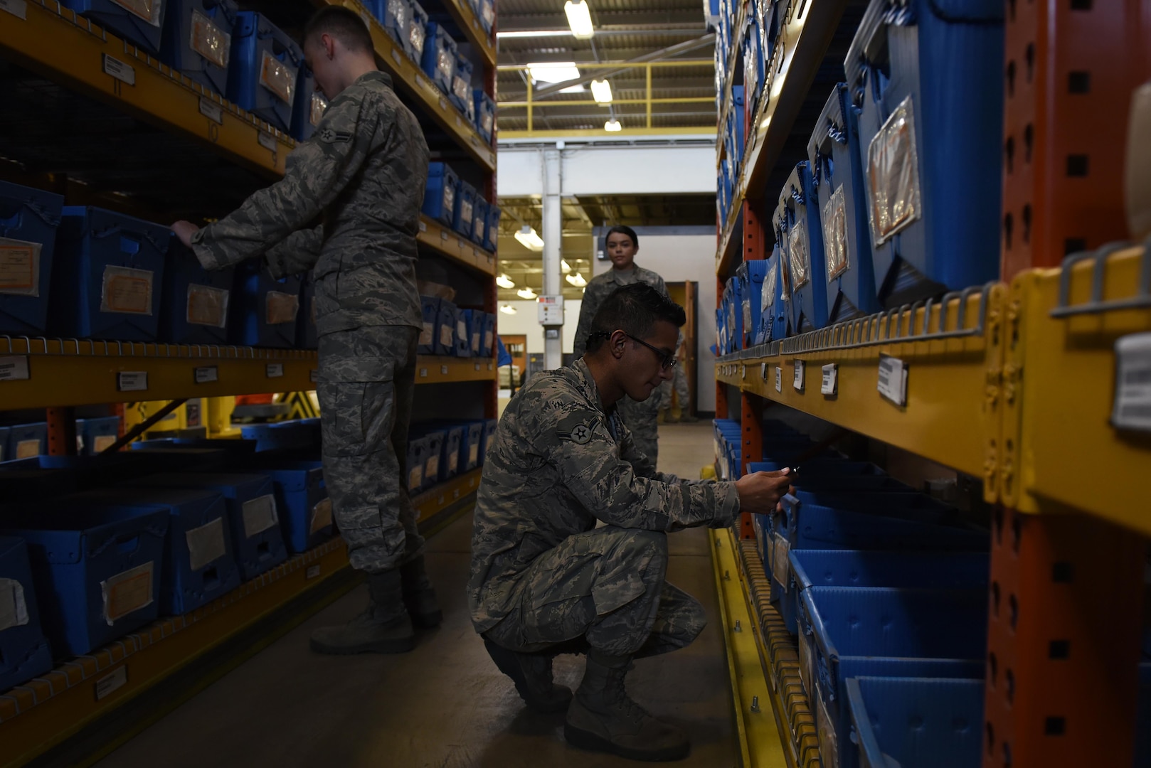 Three airmen in a warehouse search through boxes.